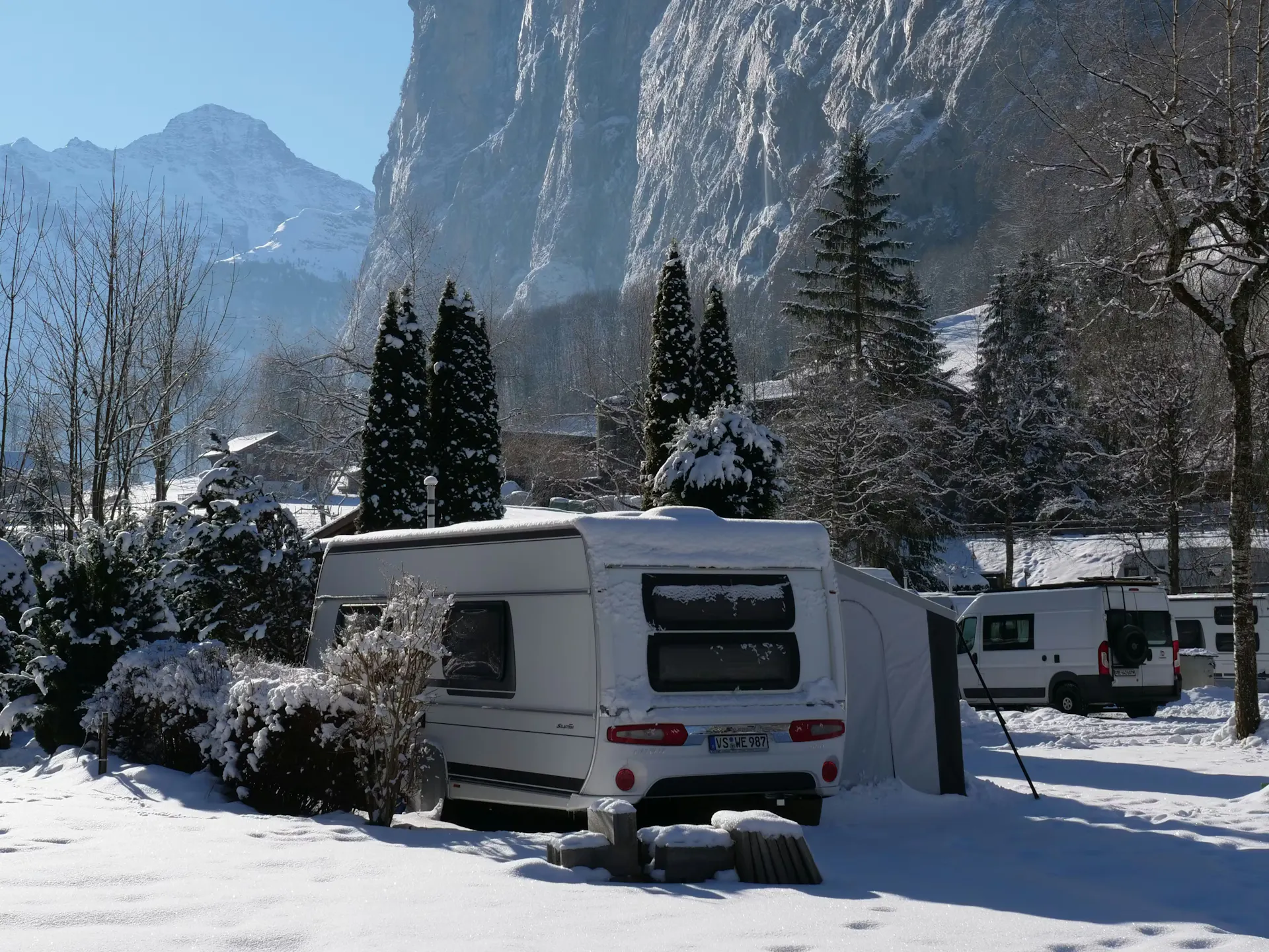 Wohnwagen im Winter auf dem Camping Jungfrau im Lauterbrunnental mit Felswänden und blauem Himmel im Hintergrund