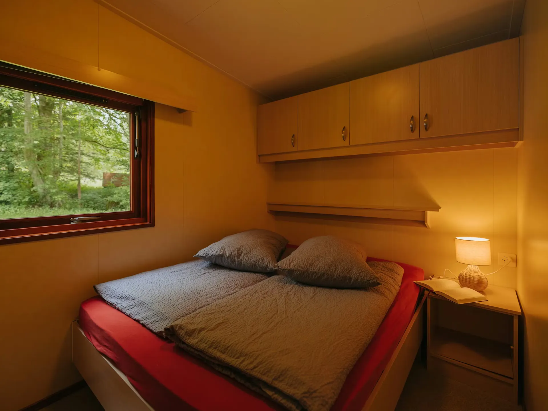 Schlafzimmer des Mobilheims Jungfrau mit einem Doppelbett, einem Nachttisch mit Beleuchtung, Schränken über dem Bett und einem Fenster an der Seite des Zimmers