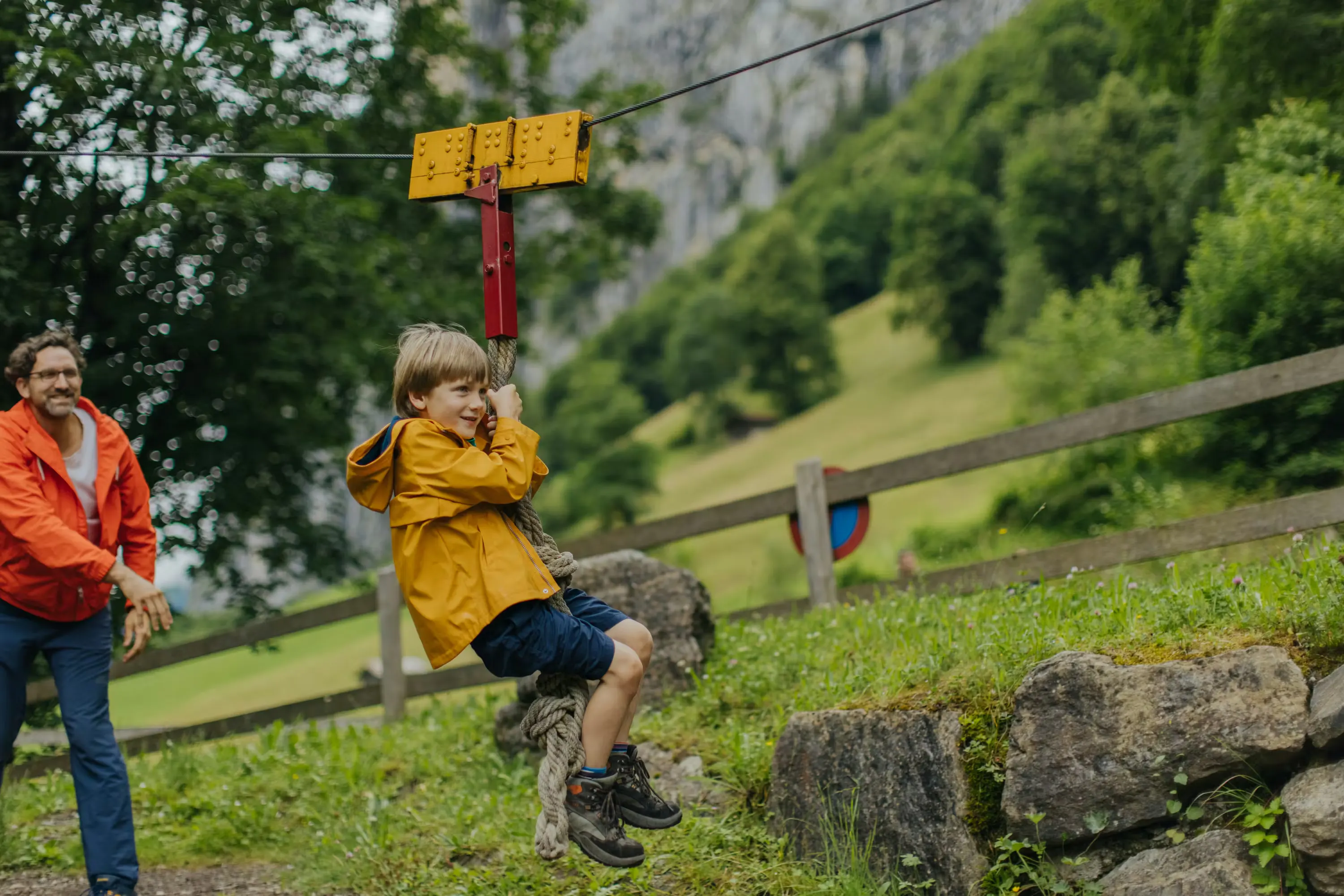 Kid on a zipline with dad helping in the background and lots of greenery around