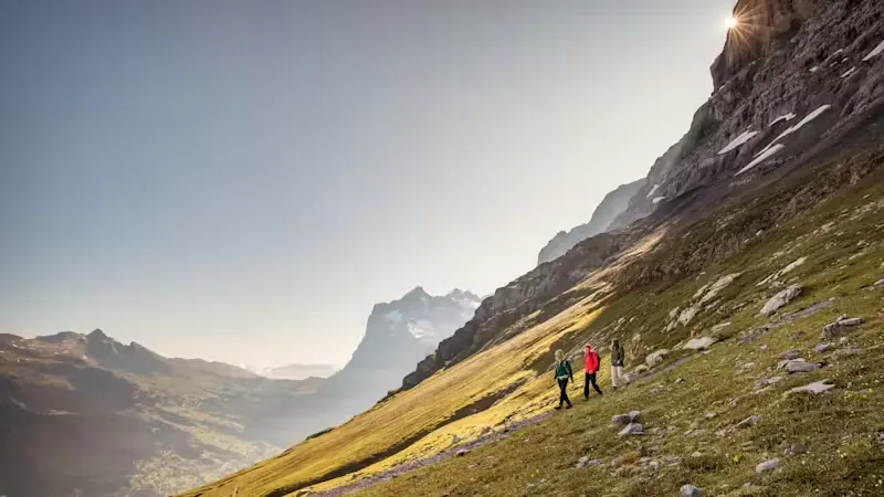 Drei Wanderer gehen einen steilen Berg hinunter, während die Sonne hinter der Felswand und den Bergen im Hintergrund untergeht