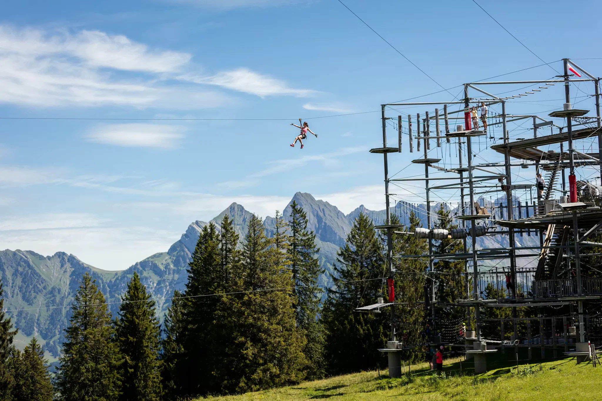 Kletterturm auf dem Flumserberg mit Kletternden und einer Frau an einer Seilrutsche. Berge im Hintergrund