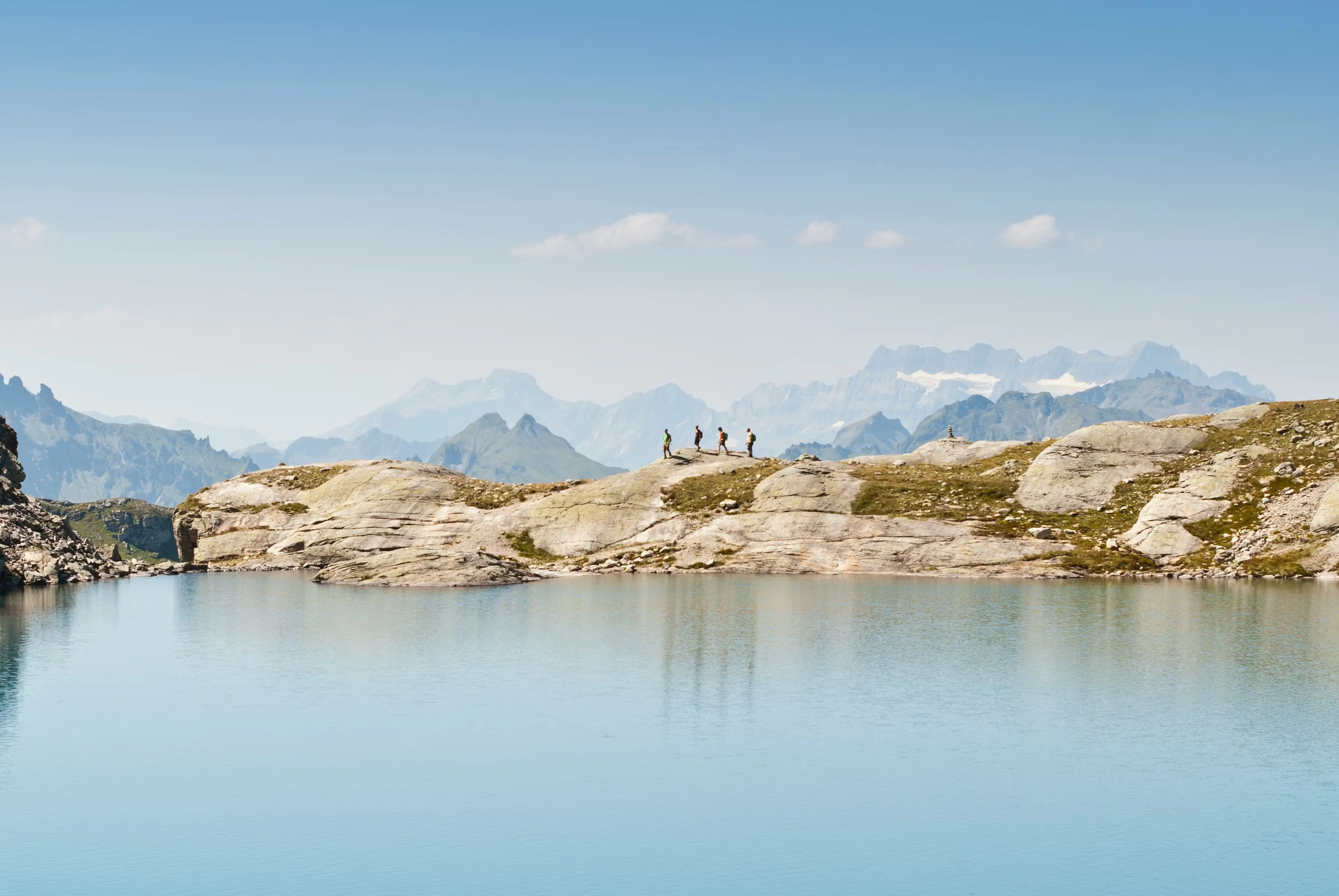 See auf der 5-Seen-Wanderung am Pizol mit vier Wanderern auf der anderen Seite des Sees auf den Felsen und hohen Bergen weit hinter ihnen