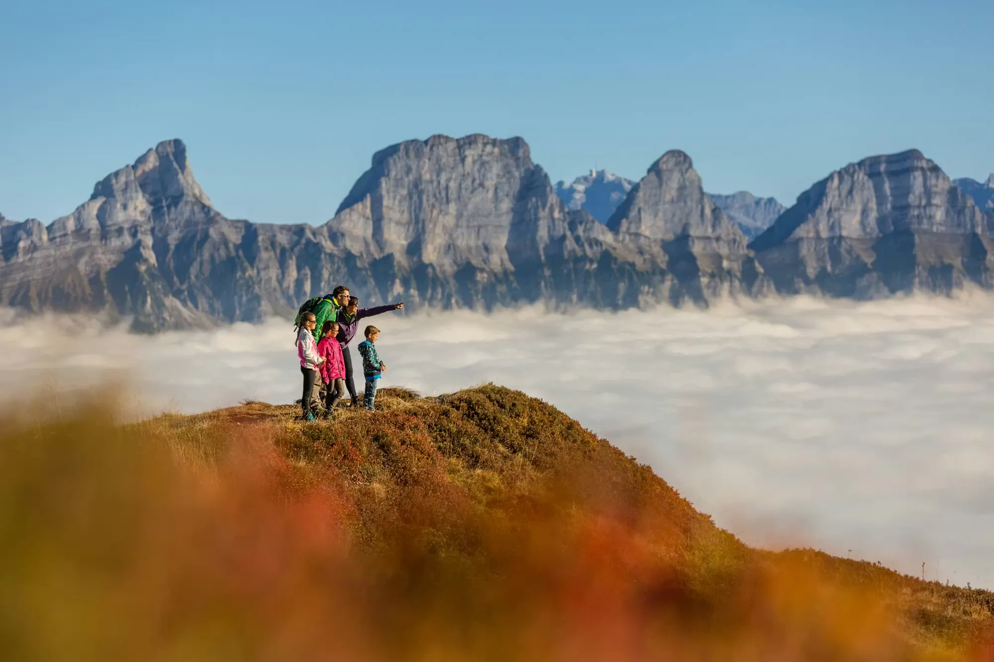 Eine Familie steht auf einem Berggipfel während einer Wanderung, unter ihnen liegt Nebel und im Hintergrund sind Berggipfel zu sehen