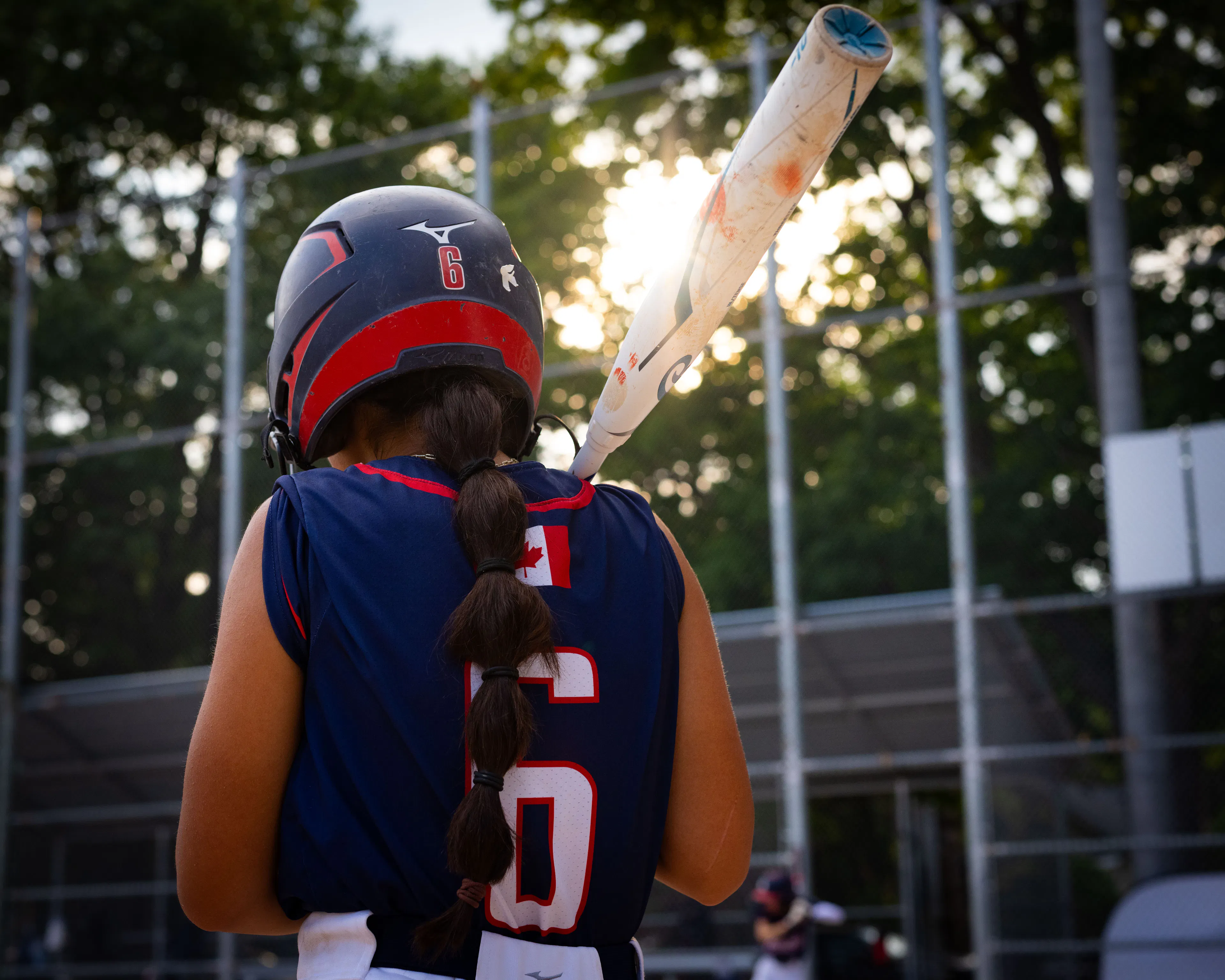 Softball player wearing helmet and jersey number 6 holding a bat, preparing to bat with the sun shining behind them