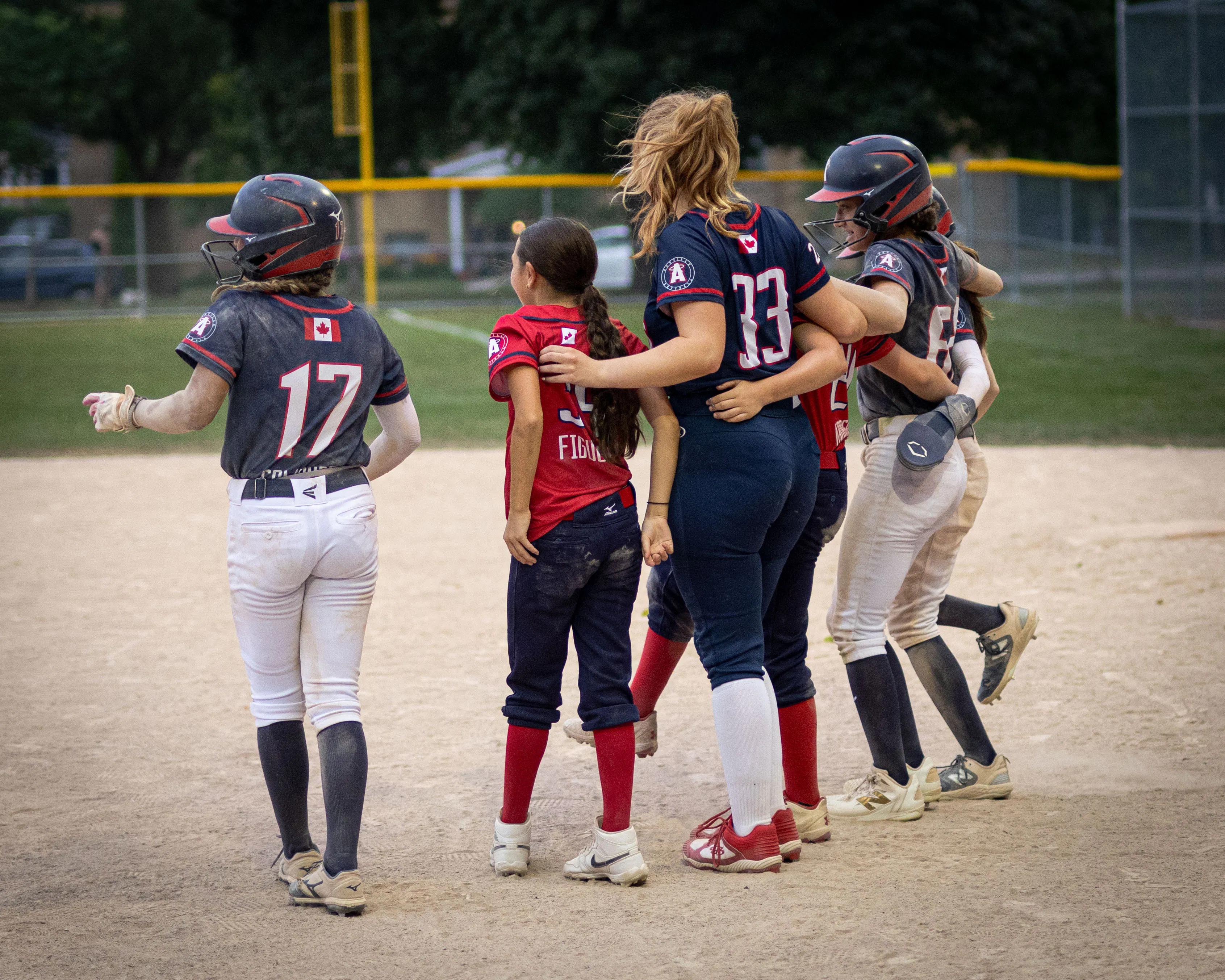 Group of softball players in red and navy uniforms with arms around each other, gathered on the infield.