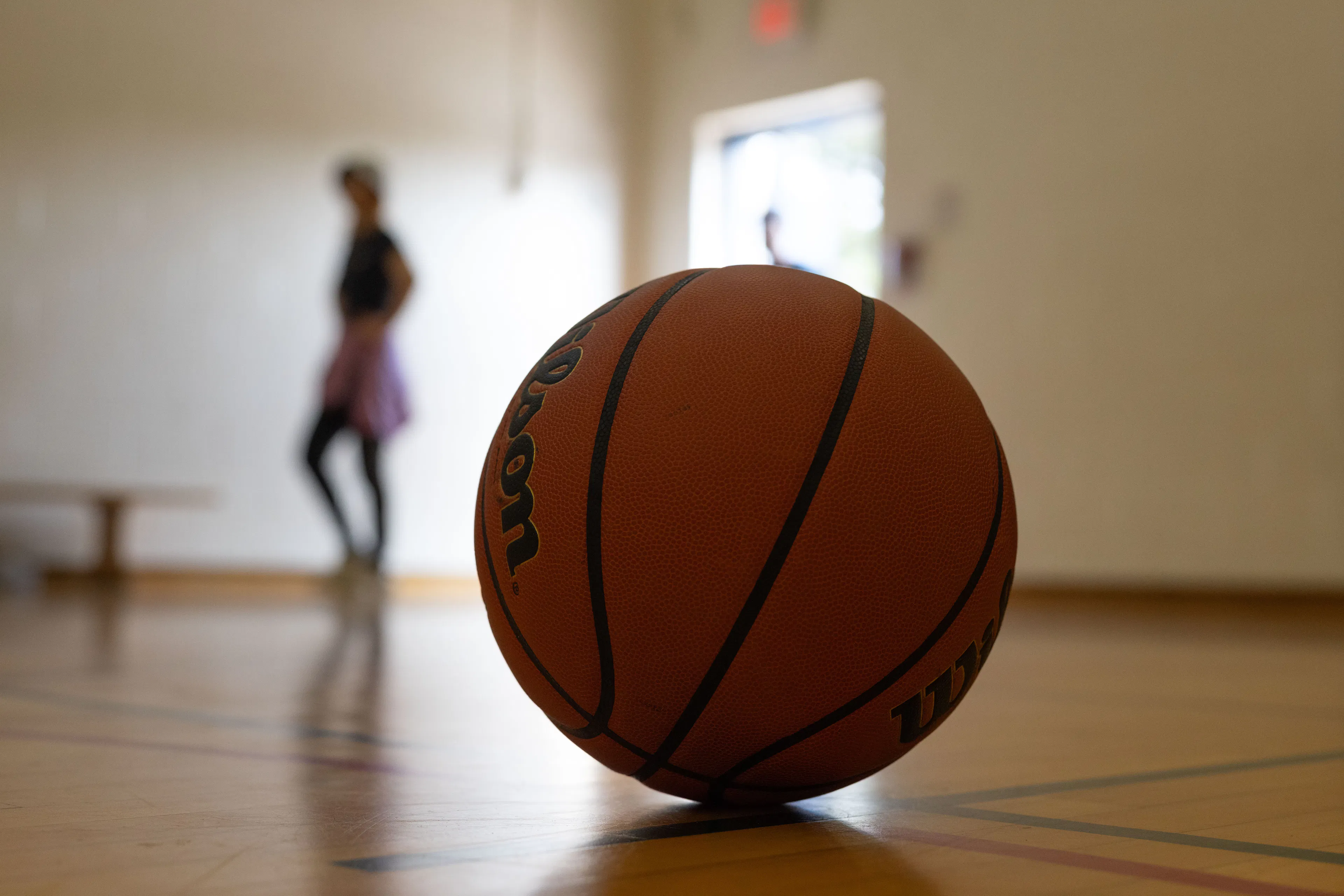 Close-up of a basketball on a wooden gym floor with a blurred figure standing near the open door in the background.