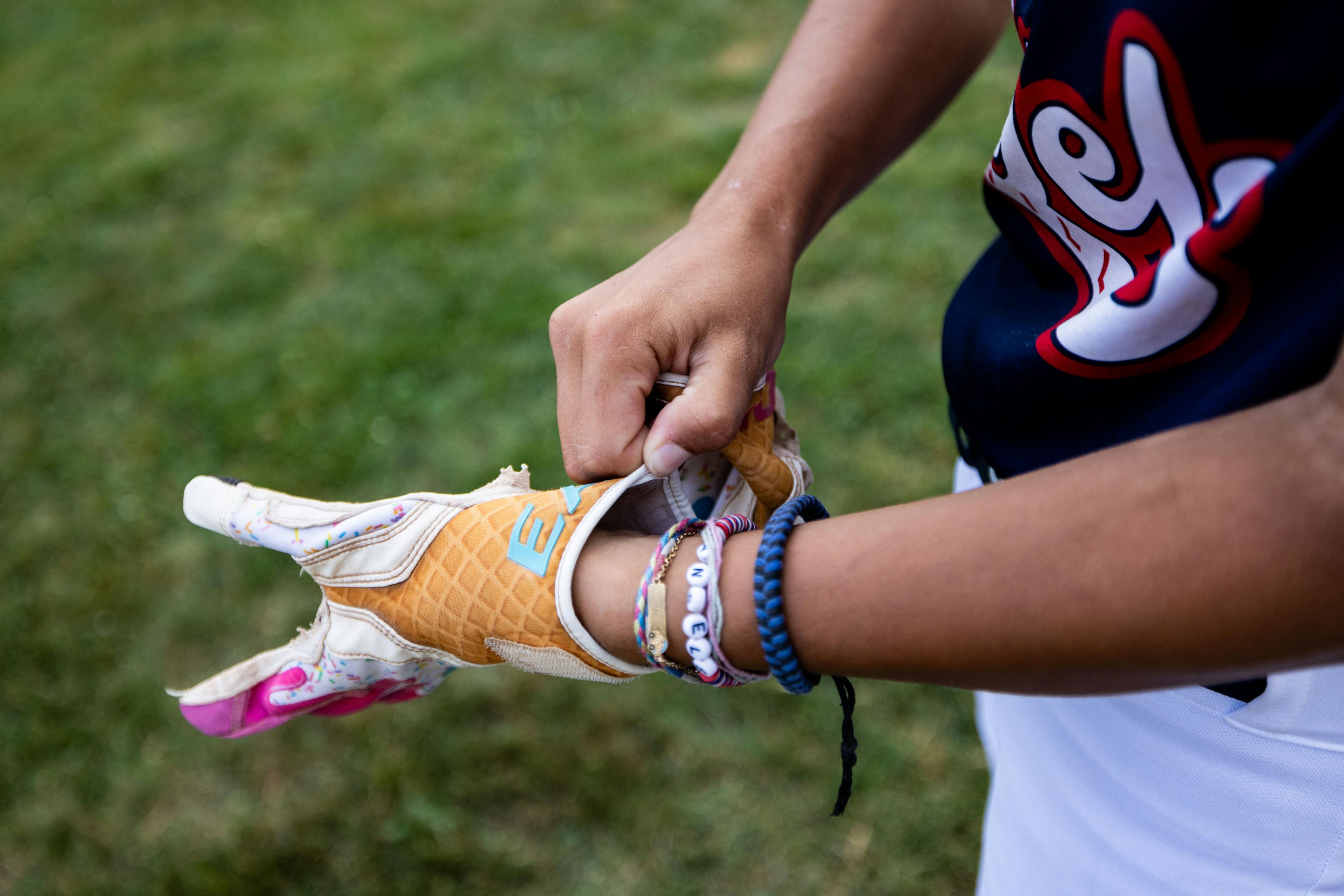Close-up of a young athlete putting on a colorful baseball batting glove, wearing friendship bracelets and a team jersey.