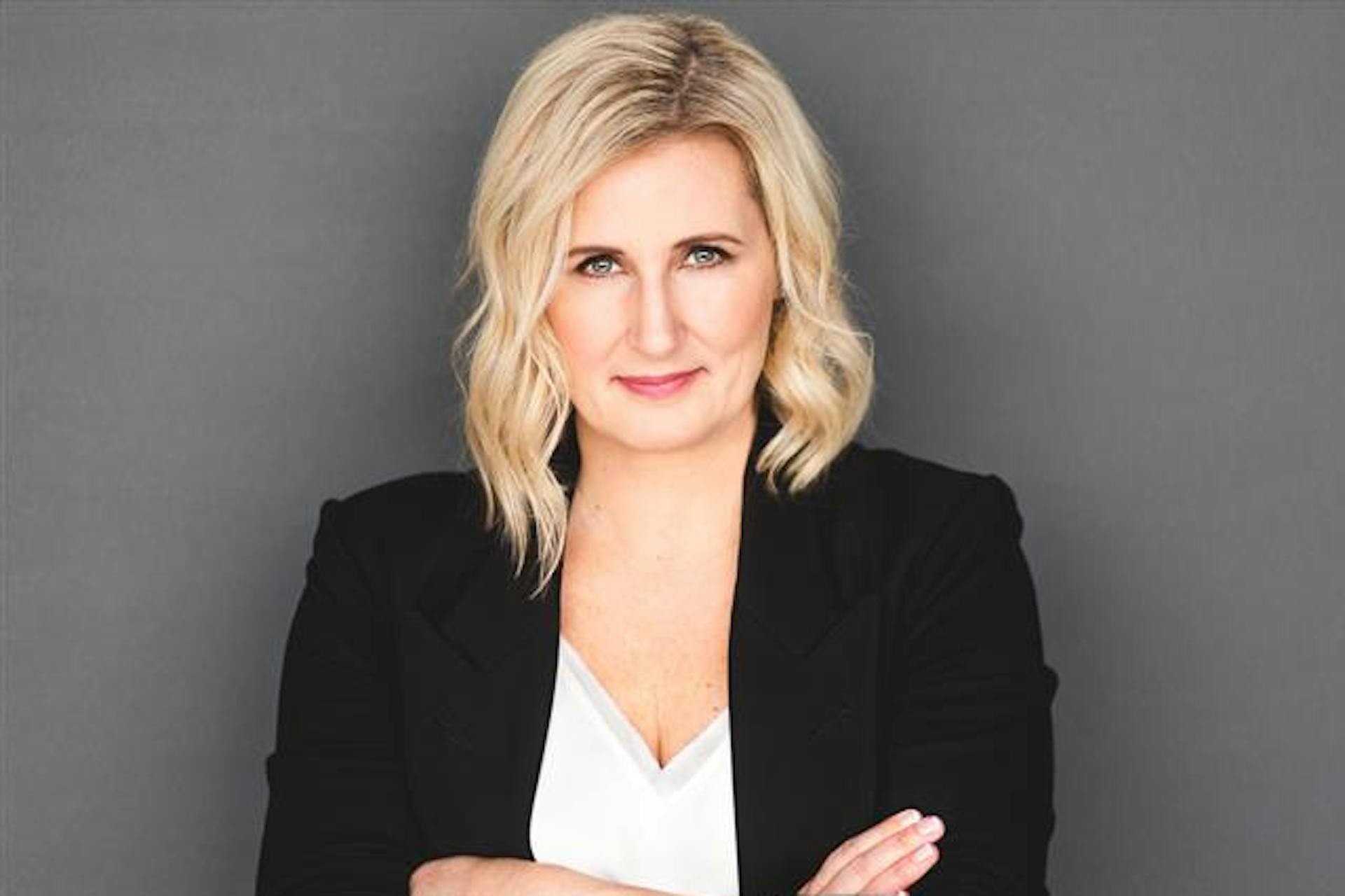 Professional portrait of a blonde woman in a black blazer and white top, arms crossed, smiling slightly against a gray background.