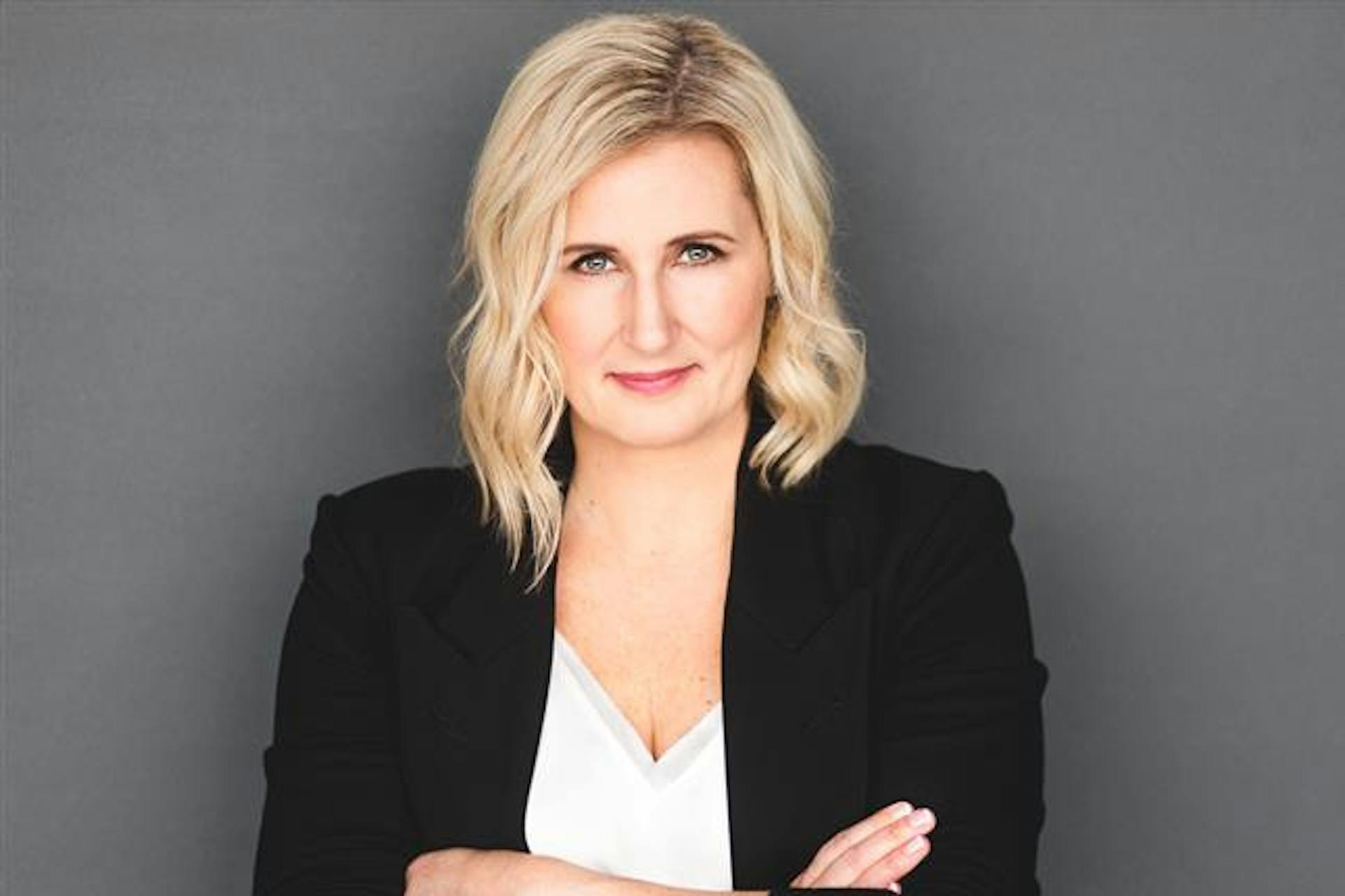 Professional portrait of a blonde woman in a black blazer and white top, arms crossed, smiling slightly against a gray background.