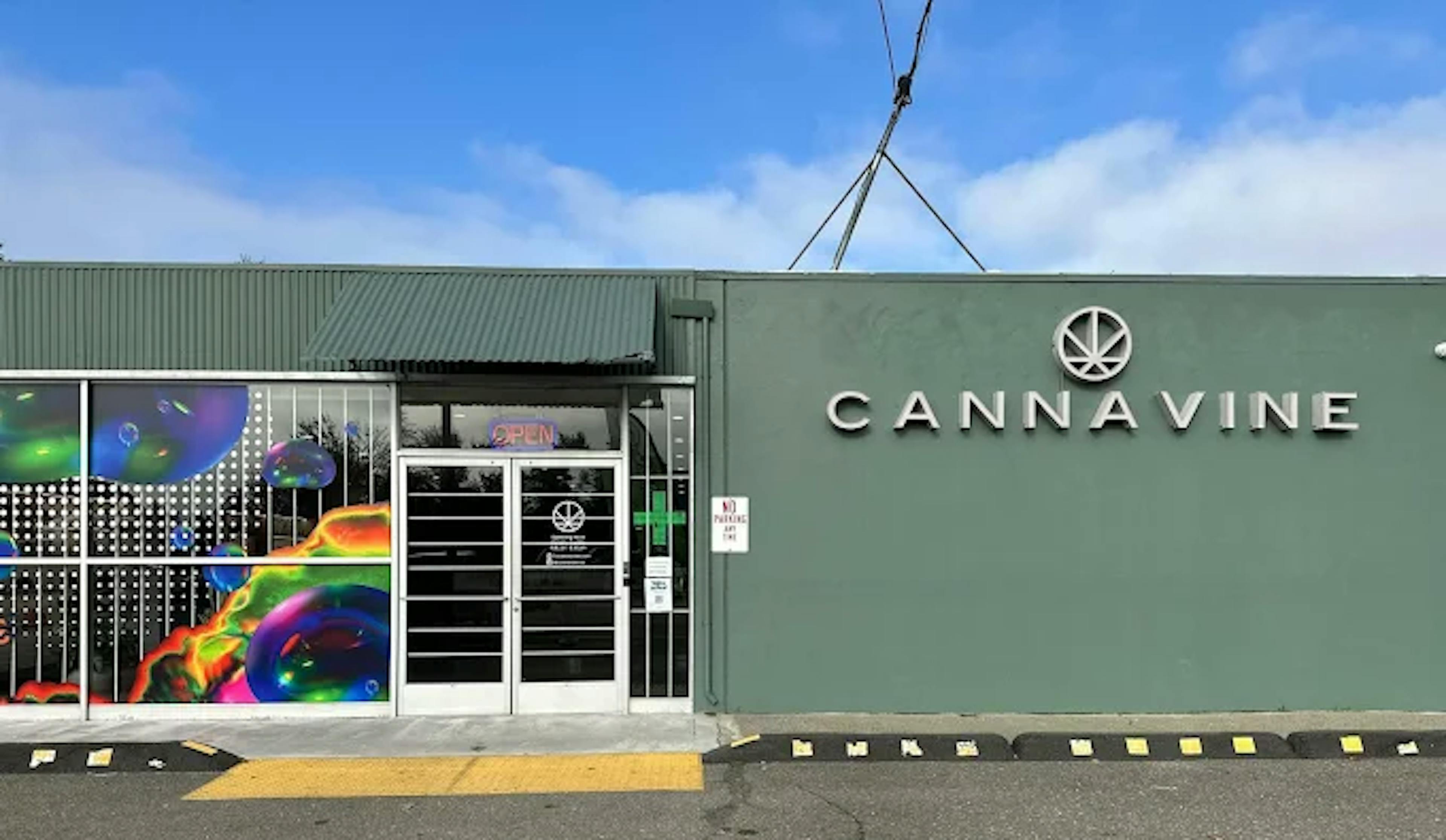 Front entrance of Cannavine Santa Rosa with metal security doors, “OPEN” signage, and colorful art glass panels. The dispensary name and logo are mounted prominently on a sage green facade.