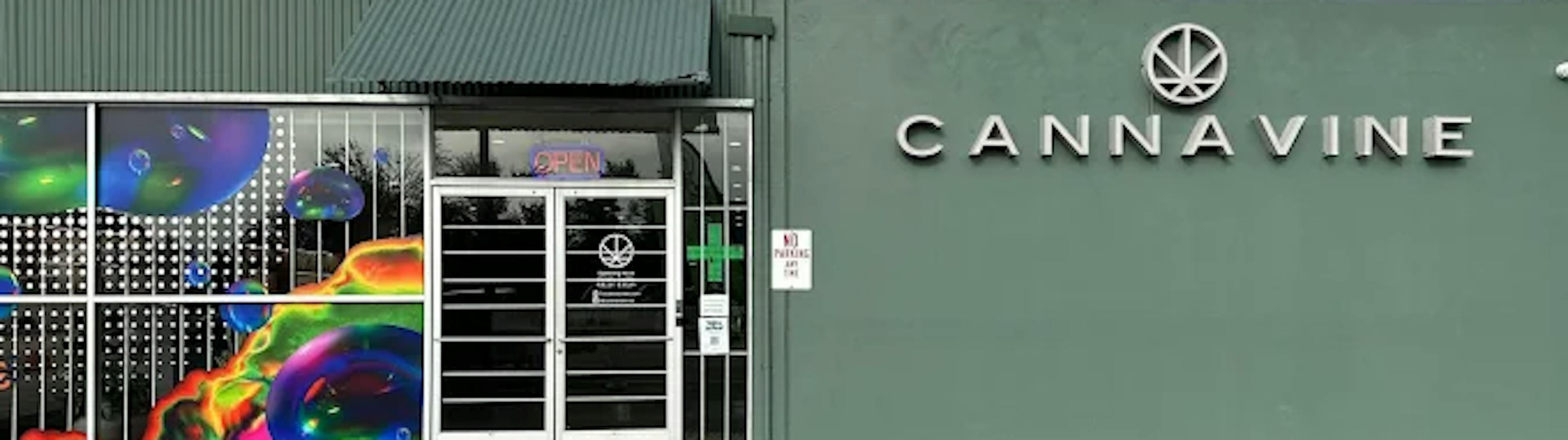 Front entrance of Cannavine Santa Rosa with metal security doors, “OPEN” signage, and colorful art glass panels. The dispensary name and logo are mounted prominently on a sage green facade.