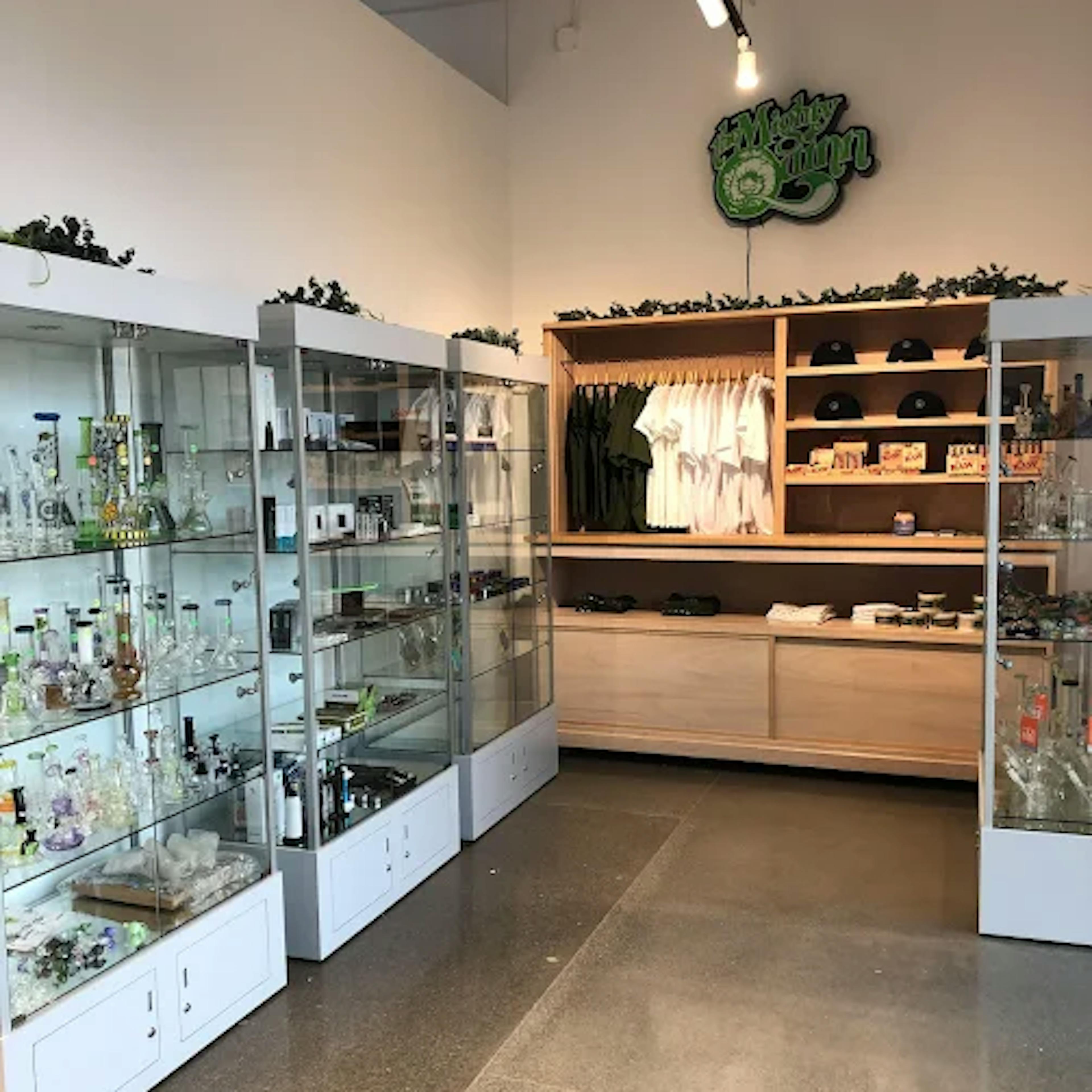 Glass cabinets filled with glassware, accessories, and smoking tools in Cannavine Ukiah. A wooden shelving unit in the back displays apparel, hats, and other branded merchandise beneath a "Muddy Green" neon-style sign.
