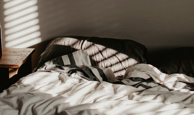 Unmade bed with crumpled white sheets and striped bedding, bathed in soft morning light coming through window blinds, casting shadow patterns on the wall and pillows.