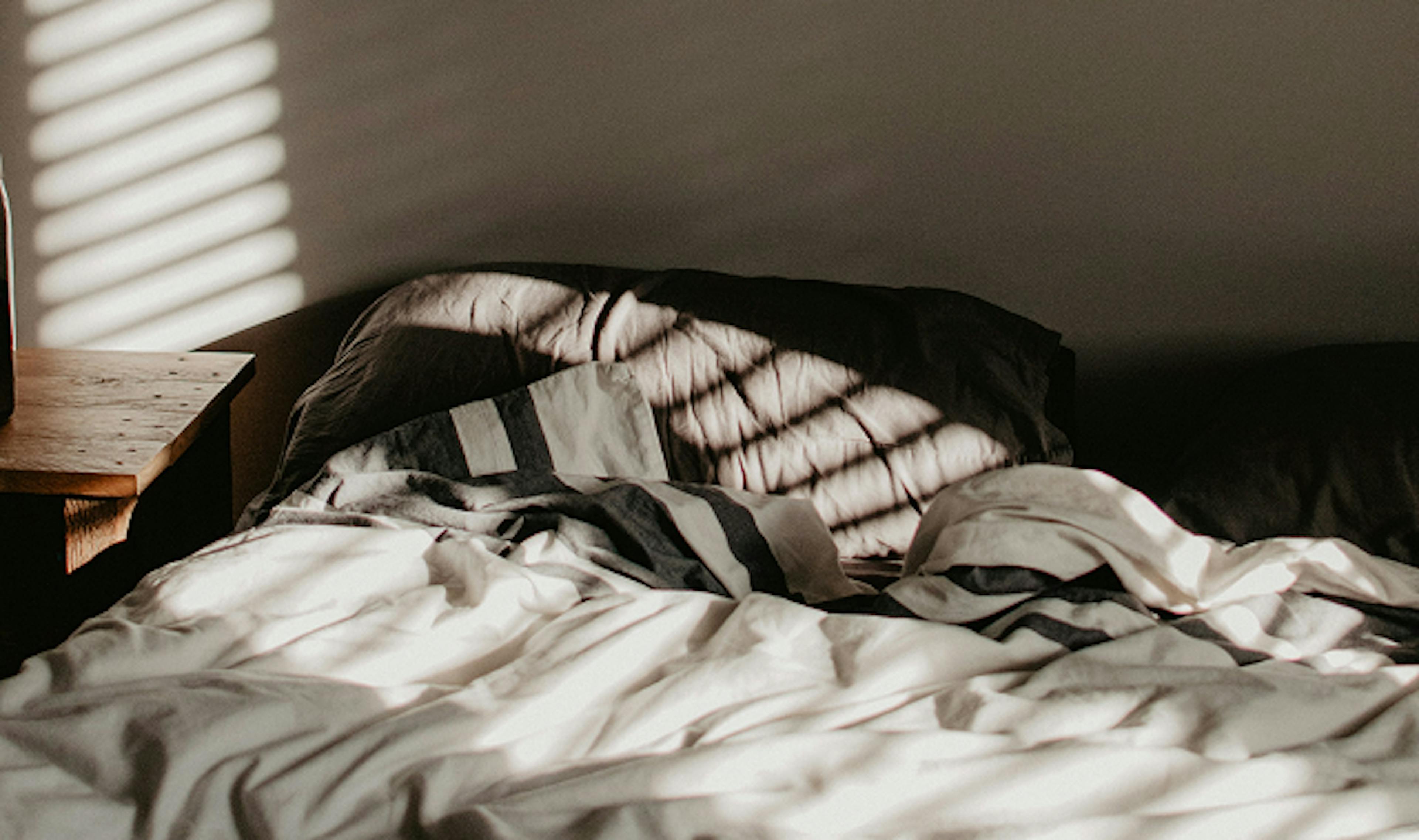 Unmade bed with crumpled white sheets and striped bedding, bathed in soft morning light coming through window blinds, casting shadow patterns on the wall and pillows.