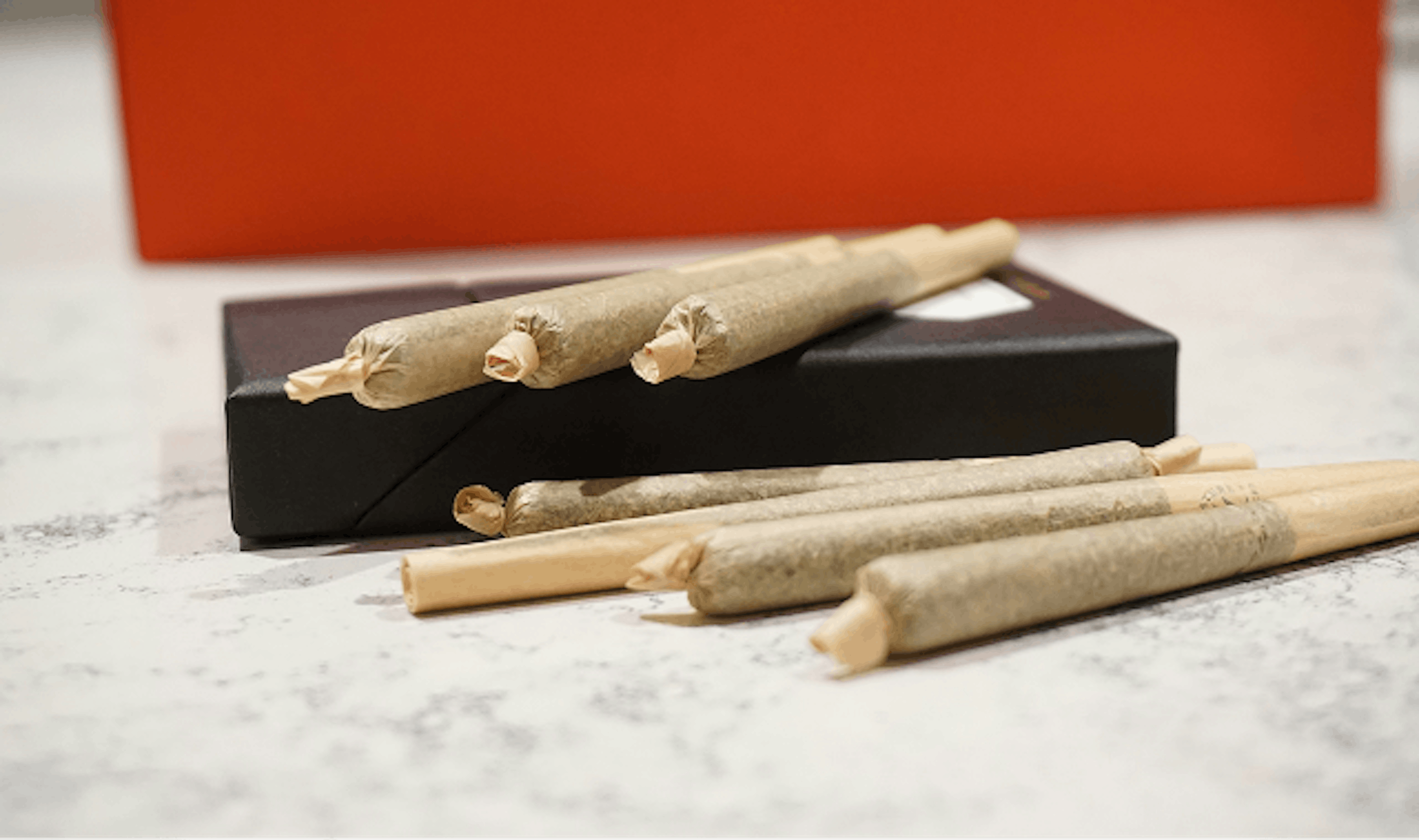 Close-up of several cannabis pre-rolls resting on a black case, with an orange background out of focus.