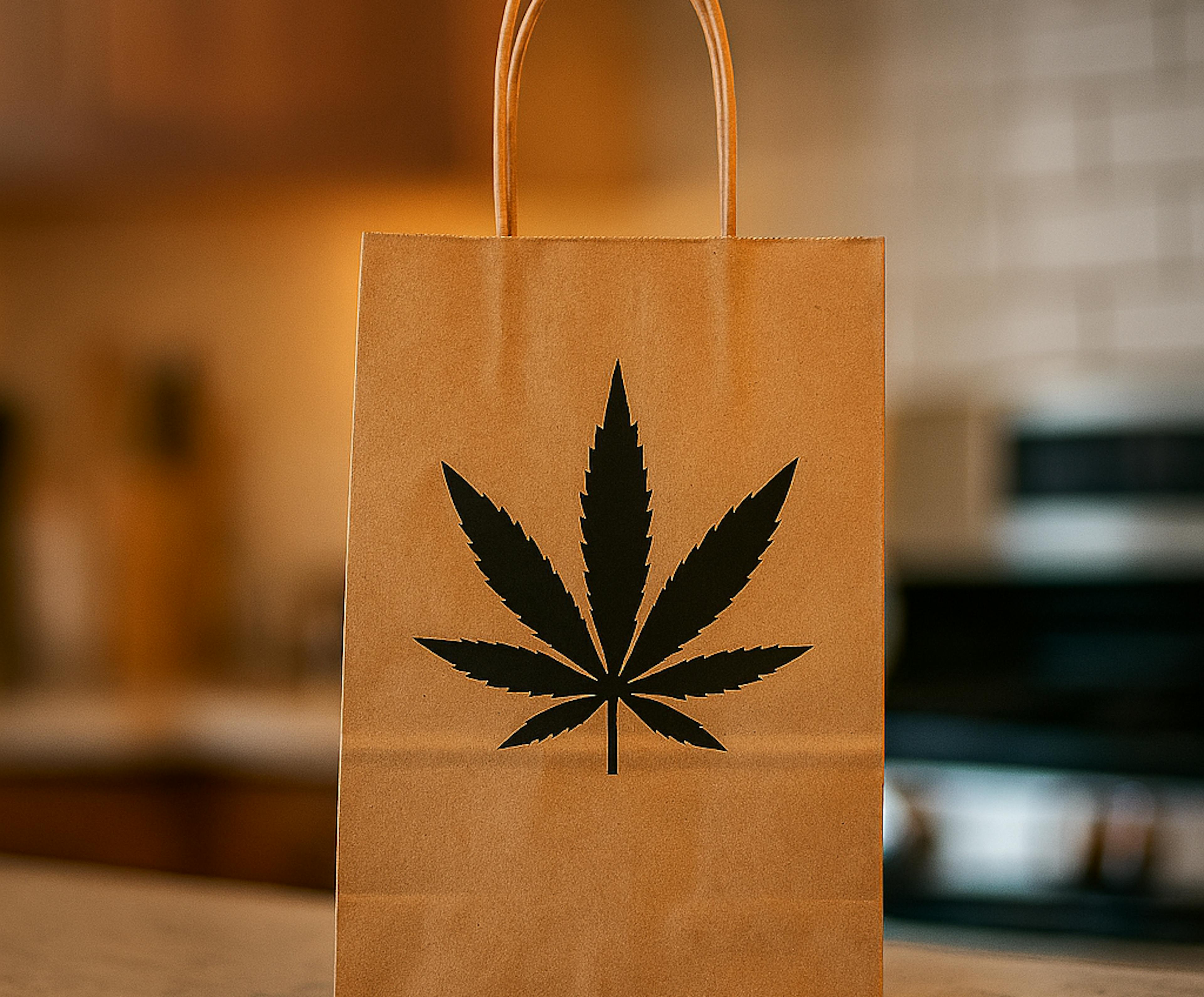 A brown paper shopping bag with a black cannabis leaf printed on the front, standing upright on a kitchen counter with a blurred stove and tile backsplash in the background.