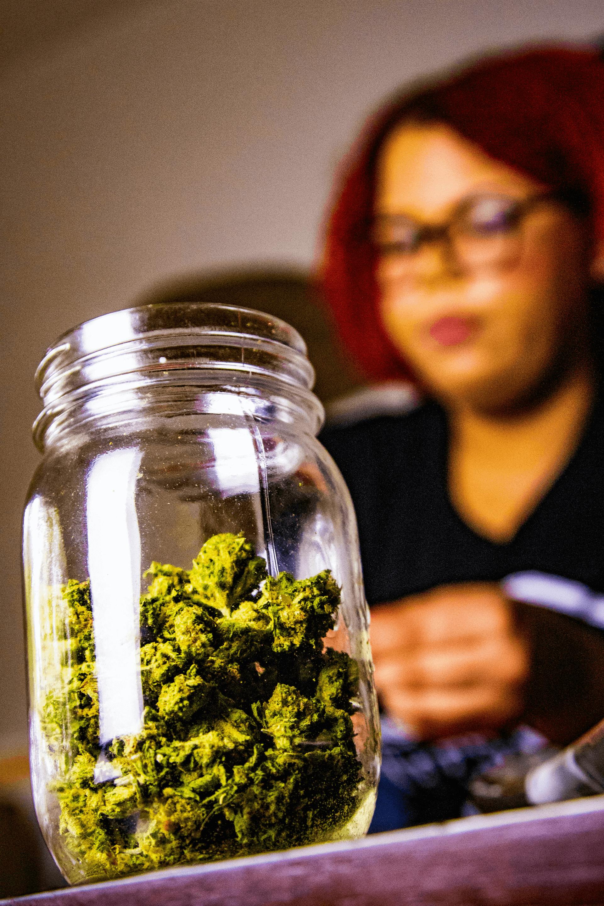 Close-up of marijuana buds in a glass jar with a blurred woman in the background