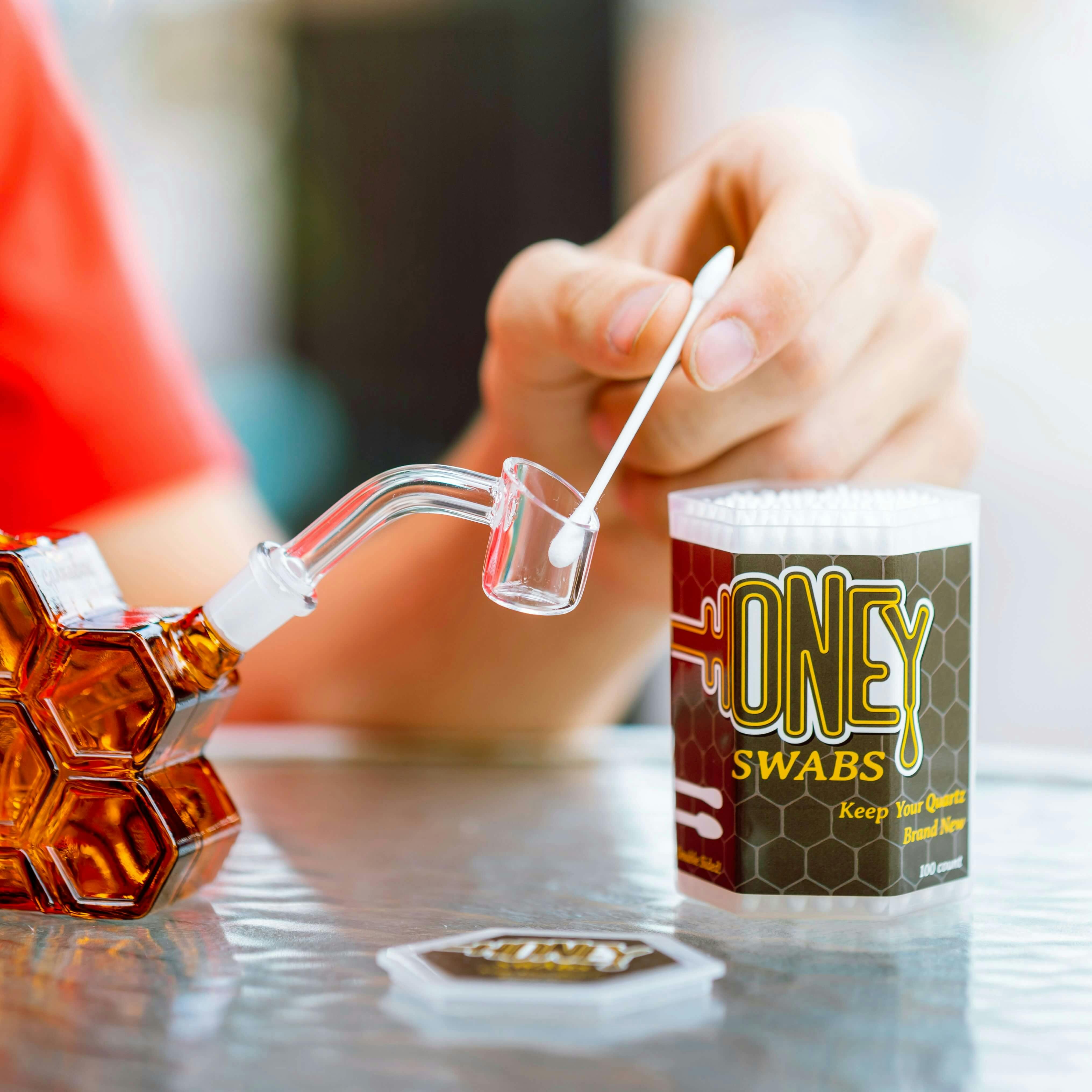 Hand cleaning a glass dab nail with a cotton swab labeled "Honey Swabs" next to a honeycomb-patterned dab rig