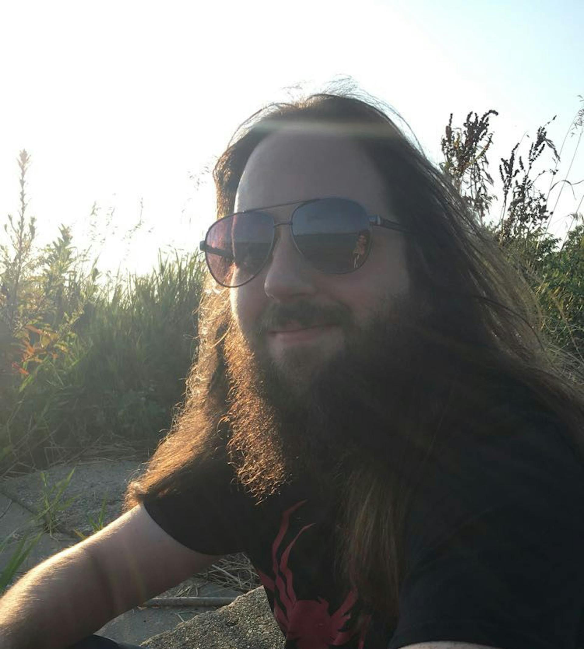 Individual with long hair, a full beard, and sunglasses, sitting outdoors in the sunlight surrounded by tall grass and plants, smiling slightly at the camera.