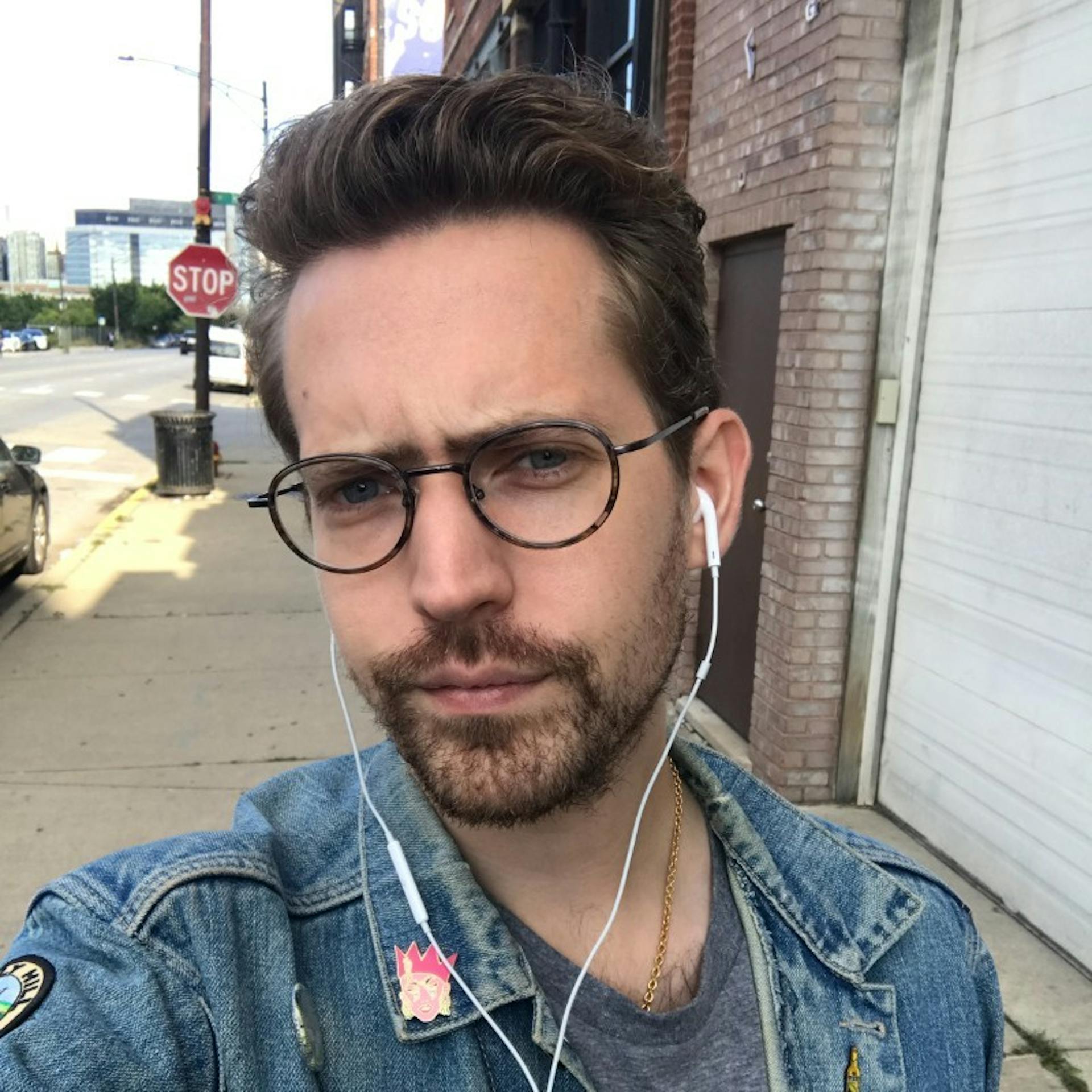 Individual with short brown hair, glasses, and a beard wearing a denim jacket and earphones, standing on a city sidewalk with a brick building and stop sign in the background.