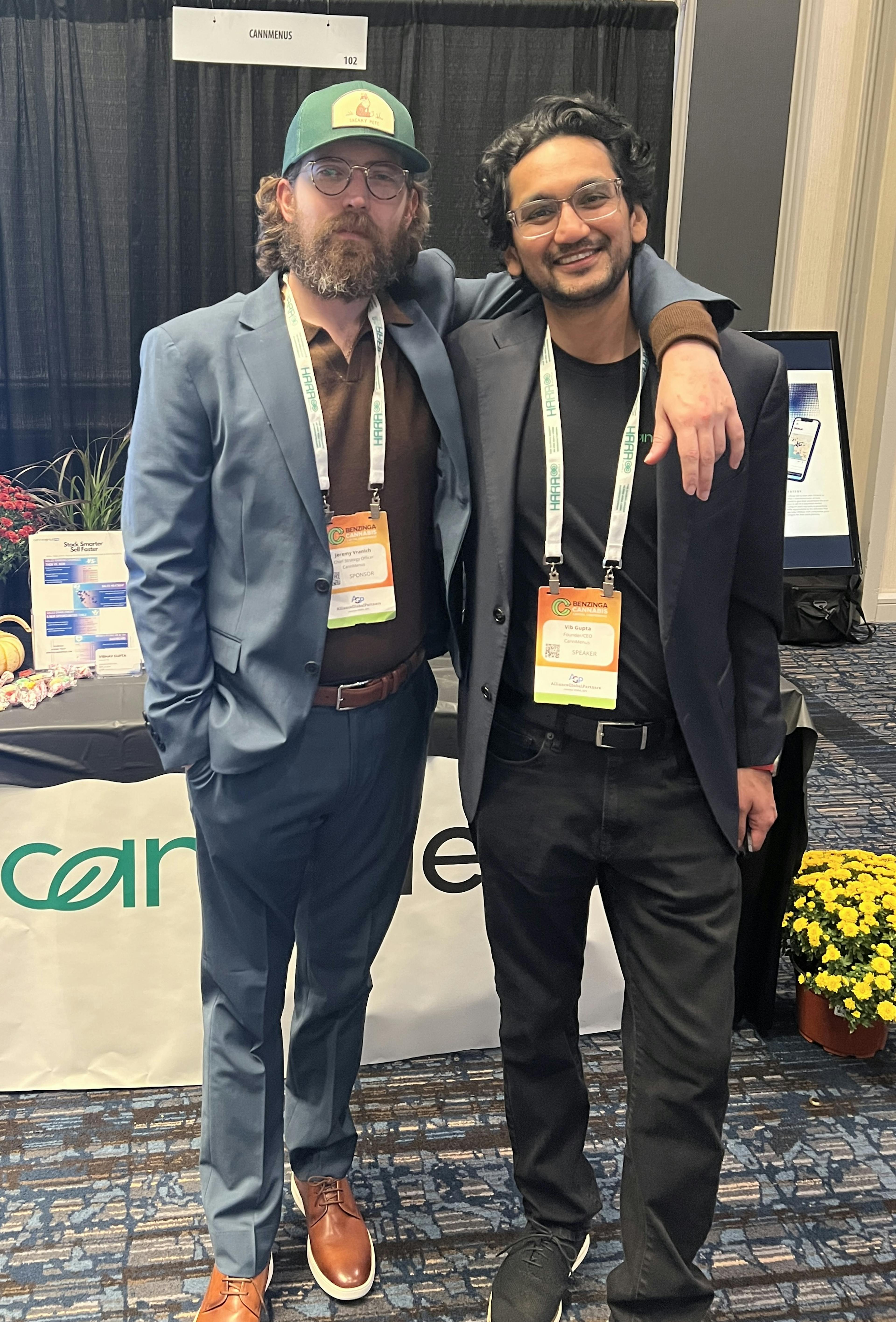 Two smiling individuals standing in front of the CannMenus booth at the Benzinga Cannabis Conference in Chicago.