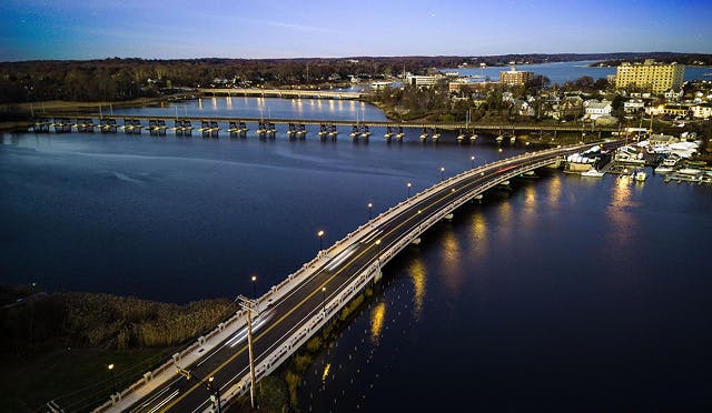 A wide lens photograph of the Navesink River estuary at sunset. The lights of the bridge are reflecting off the water and the sky is a mixture of purple and blue.
