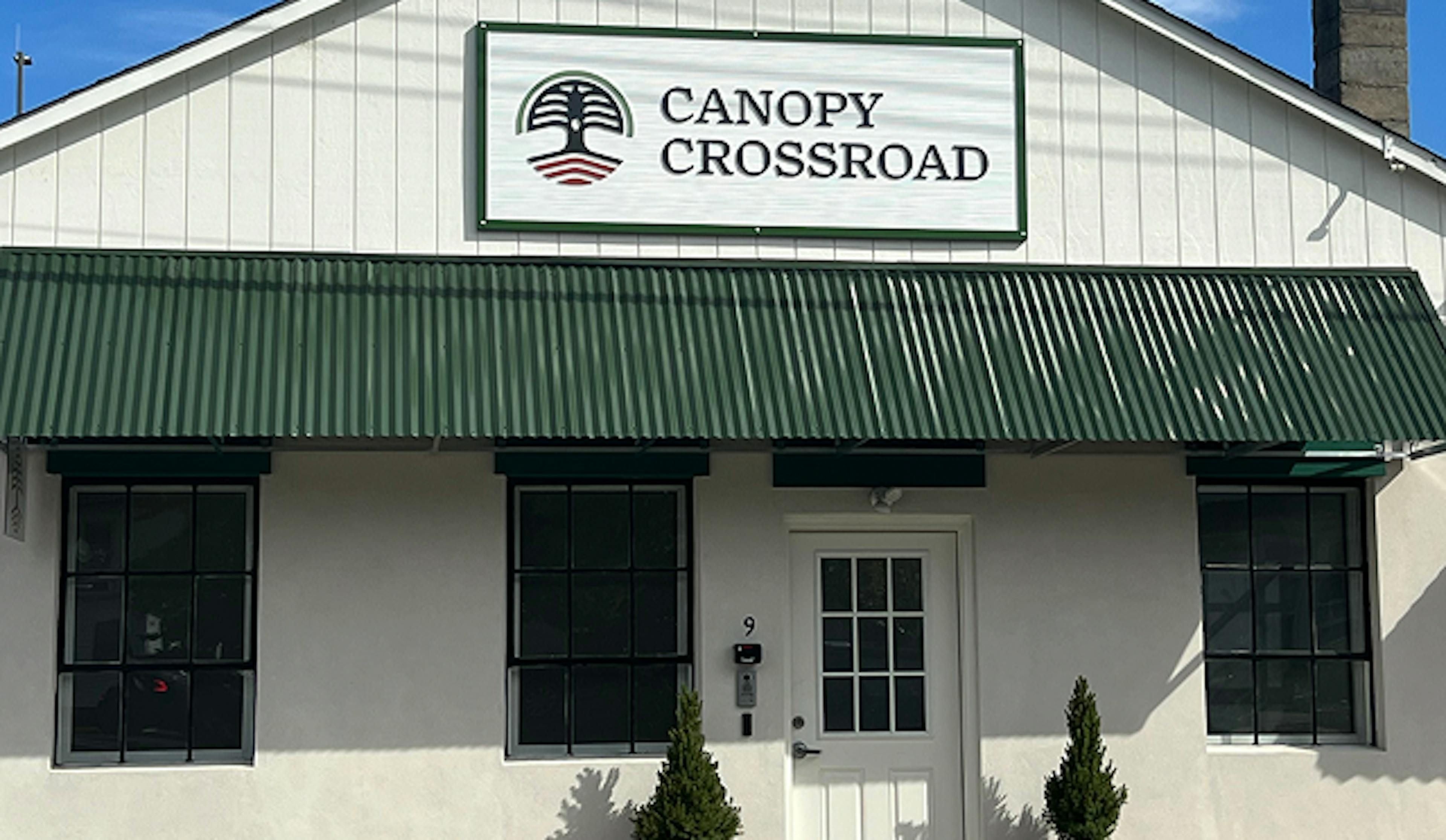 A sunny photograph of the Canopy Crossroad storefront, a white building with a green canopy. There are two potted plants outside the front door.