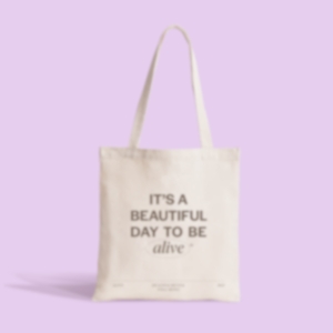 A tote bag with the text 'it's a beautiful day to be alive' against a pink background.