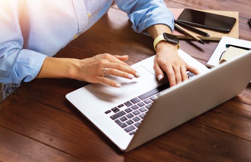 Person navigating on a laptop computer sitting at a table with their phone, notepad and writing utensils next to them.