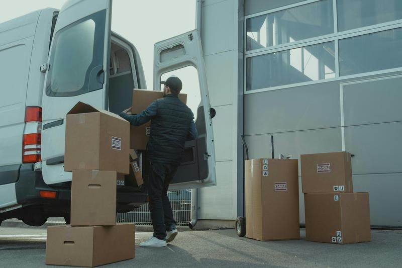 Person loads cardboard boxes into a white delivery van outside a warehouse. Boxes labeled "FRAGILE".