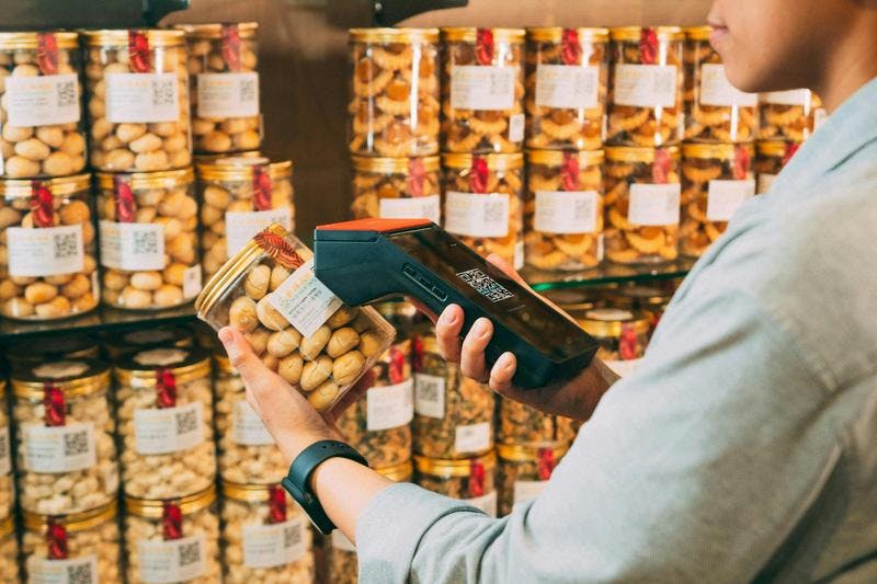 Person scans jar of snacks with barcode reader in store, surrounded by shelves filled with similar jars.