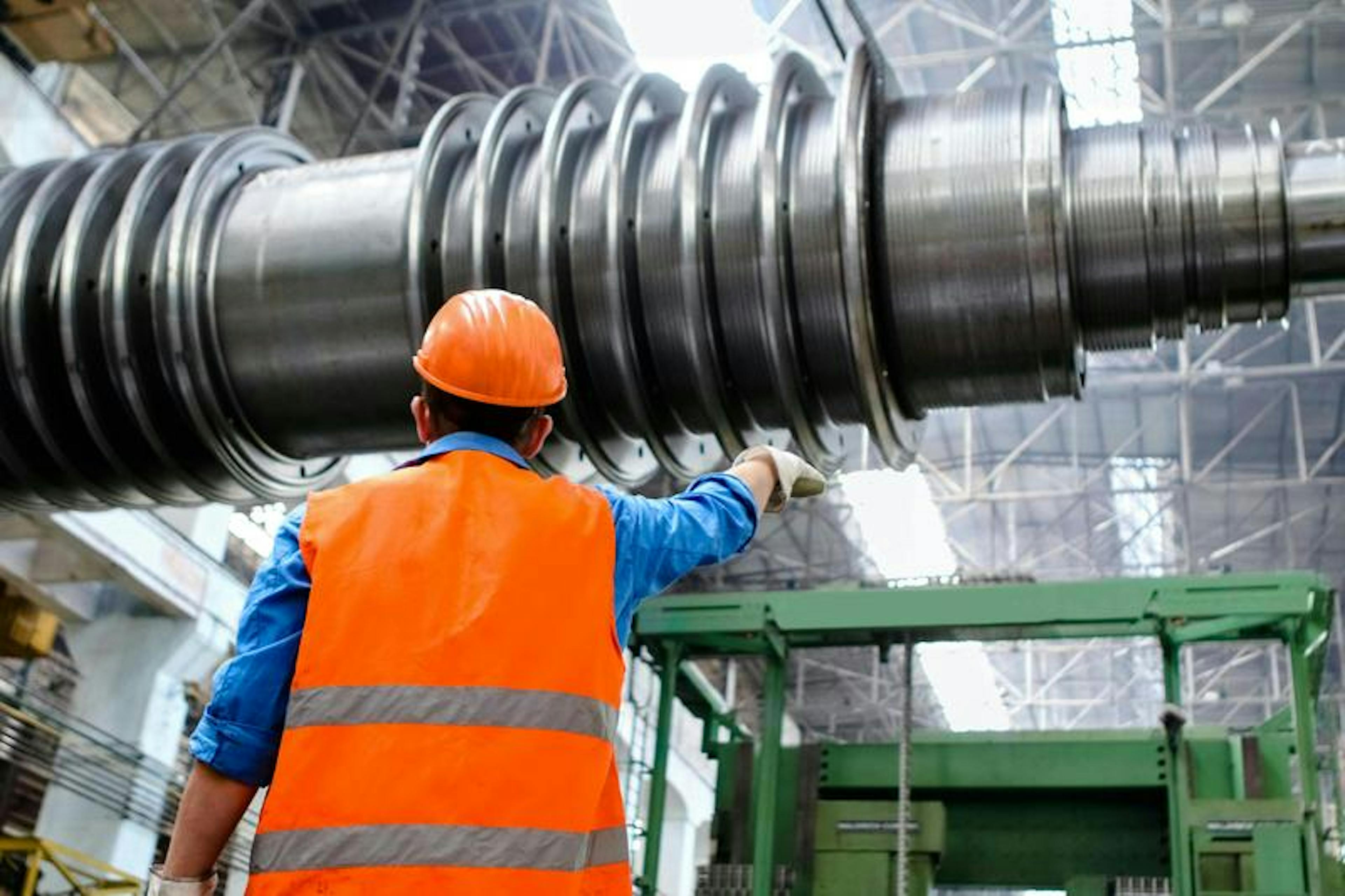 Worker in orange vest and helmet points at large industrial machine in a spacious factory.