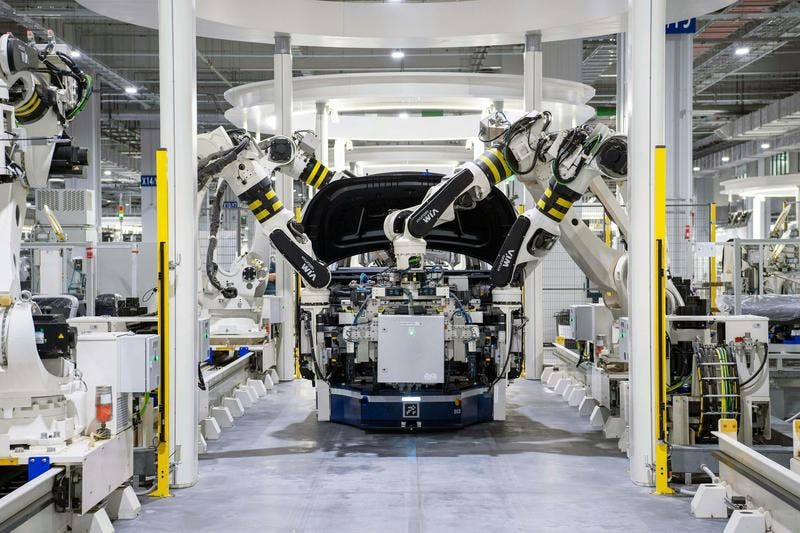 Robotic arms assemble a car in a factory with surrounding machinery and control panels.