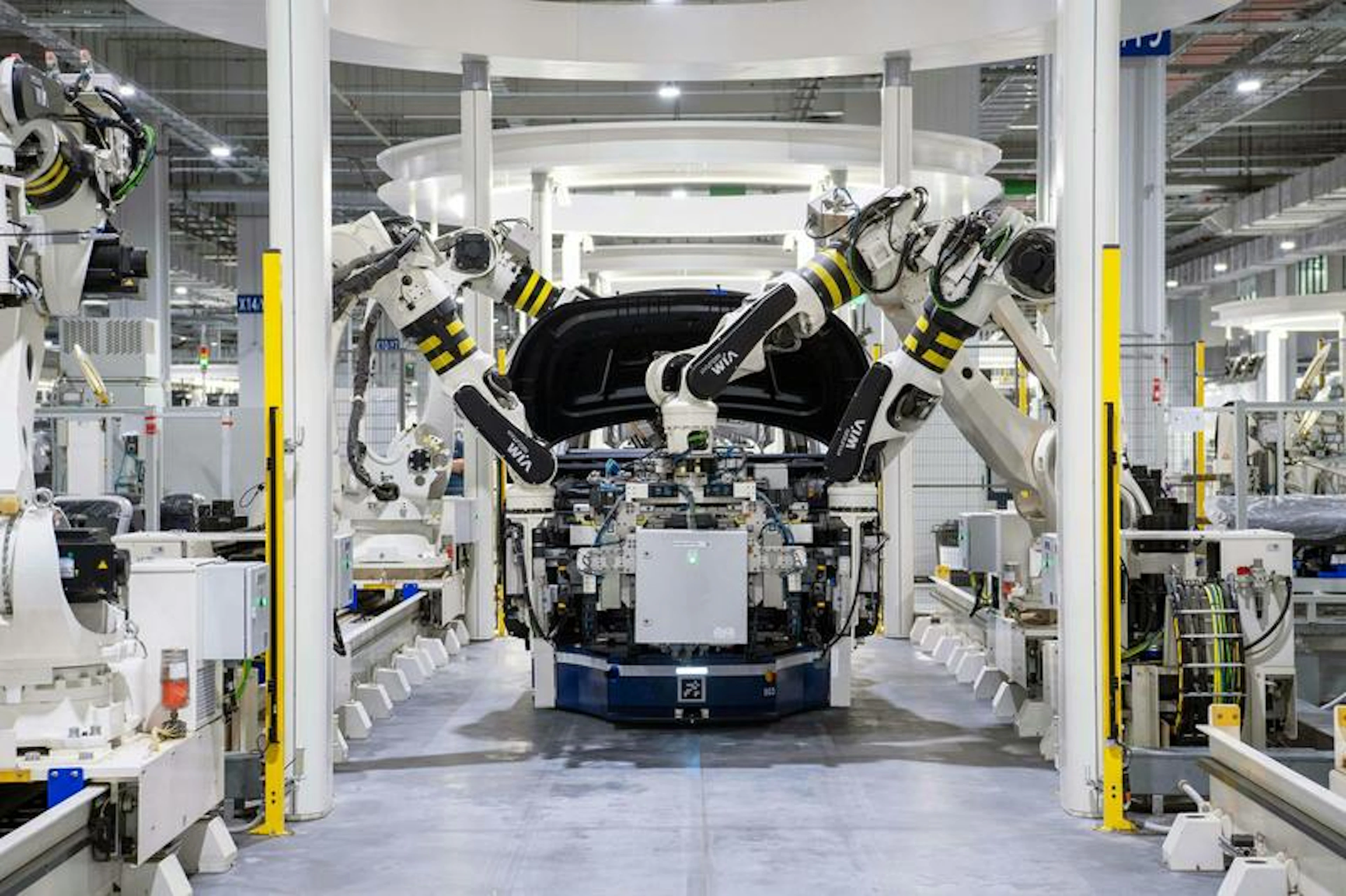 Robotic arms assemble a car in a factory with surrounding machinery and control panels.