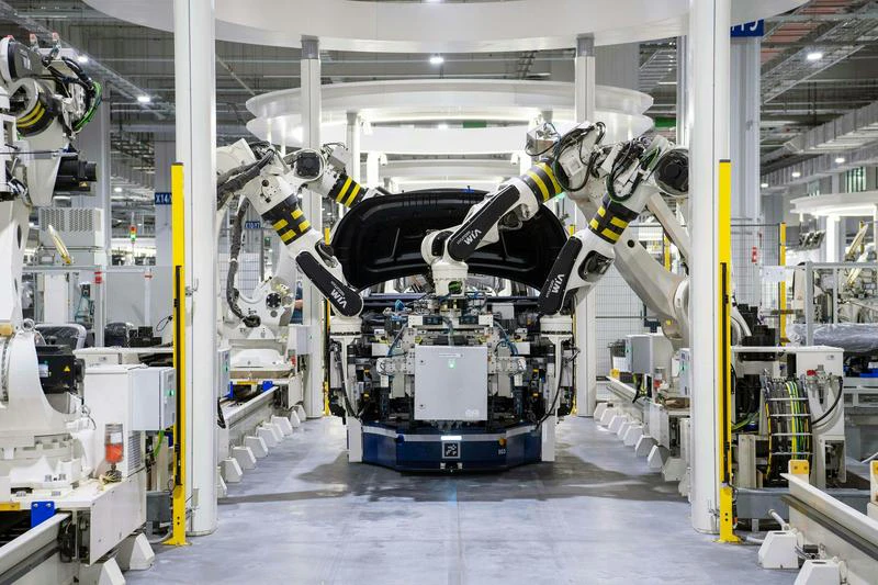 Robotic arms assemble a car in a factory with surrounding machinery and control panels.