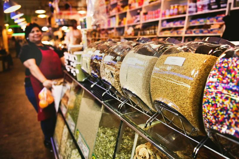 Glass jars of grains and candies on market counter; vendor in red apron with bags behind shelves.