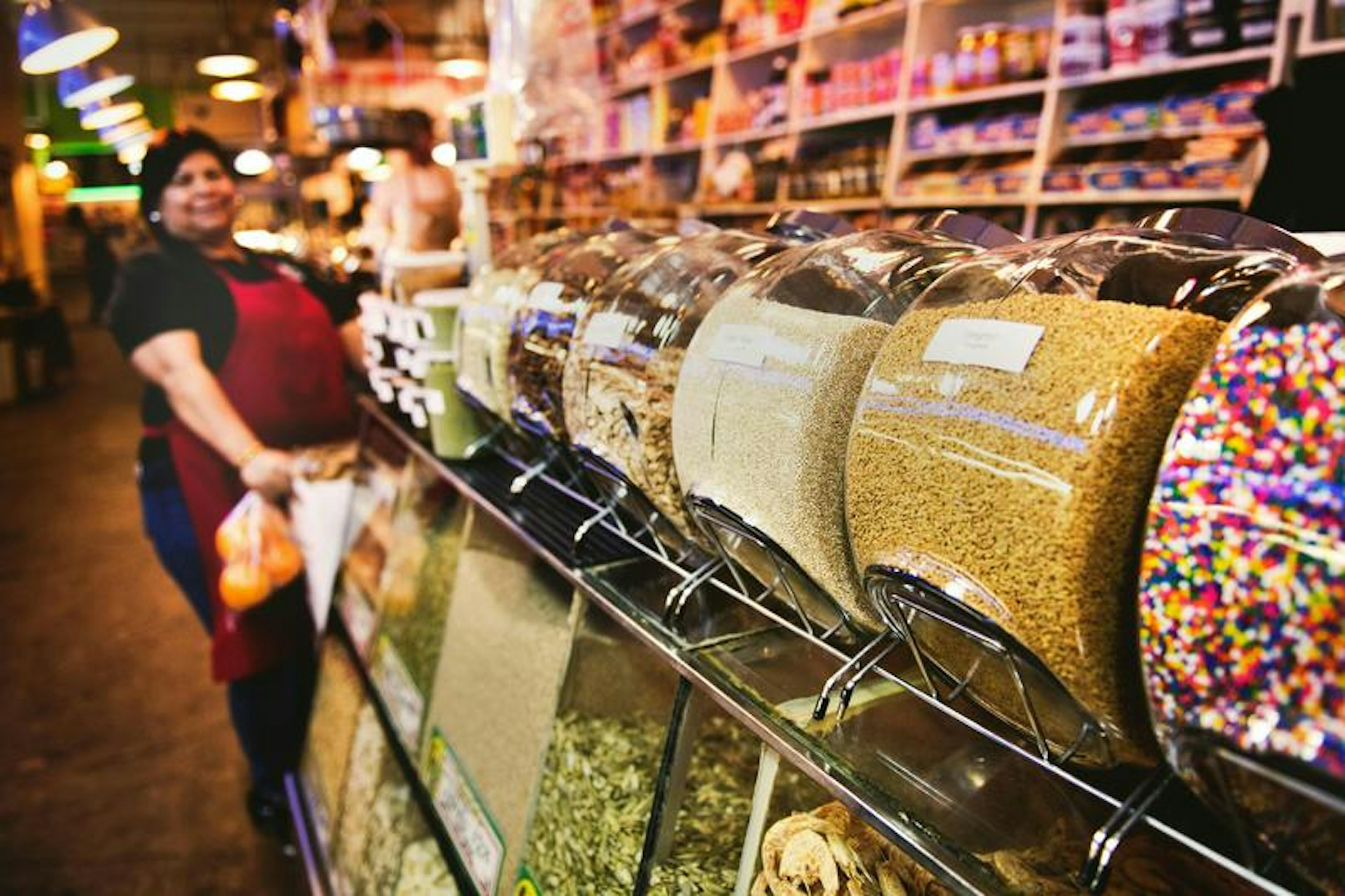 Glass jars of grains and candies on market counter; vendor in red apron with bags behind shelves.