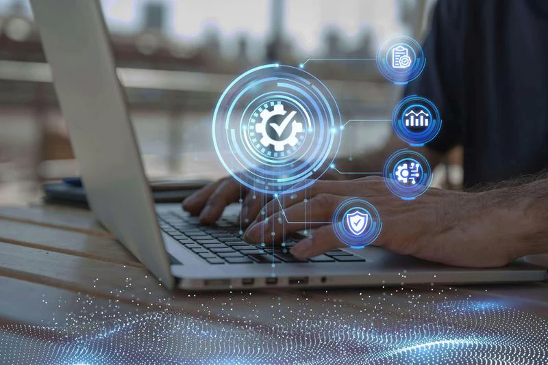 Hands type on a laptop, overlaid with digital icons: gear, checkmark, chart, shield. Outdoor scene, blurred cityscape.