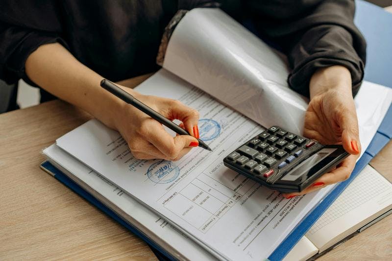 Person writes on documents with a pen while holding a calculator on a wooden desk in an office.