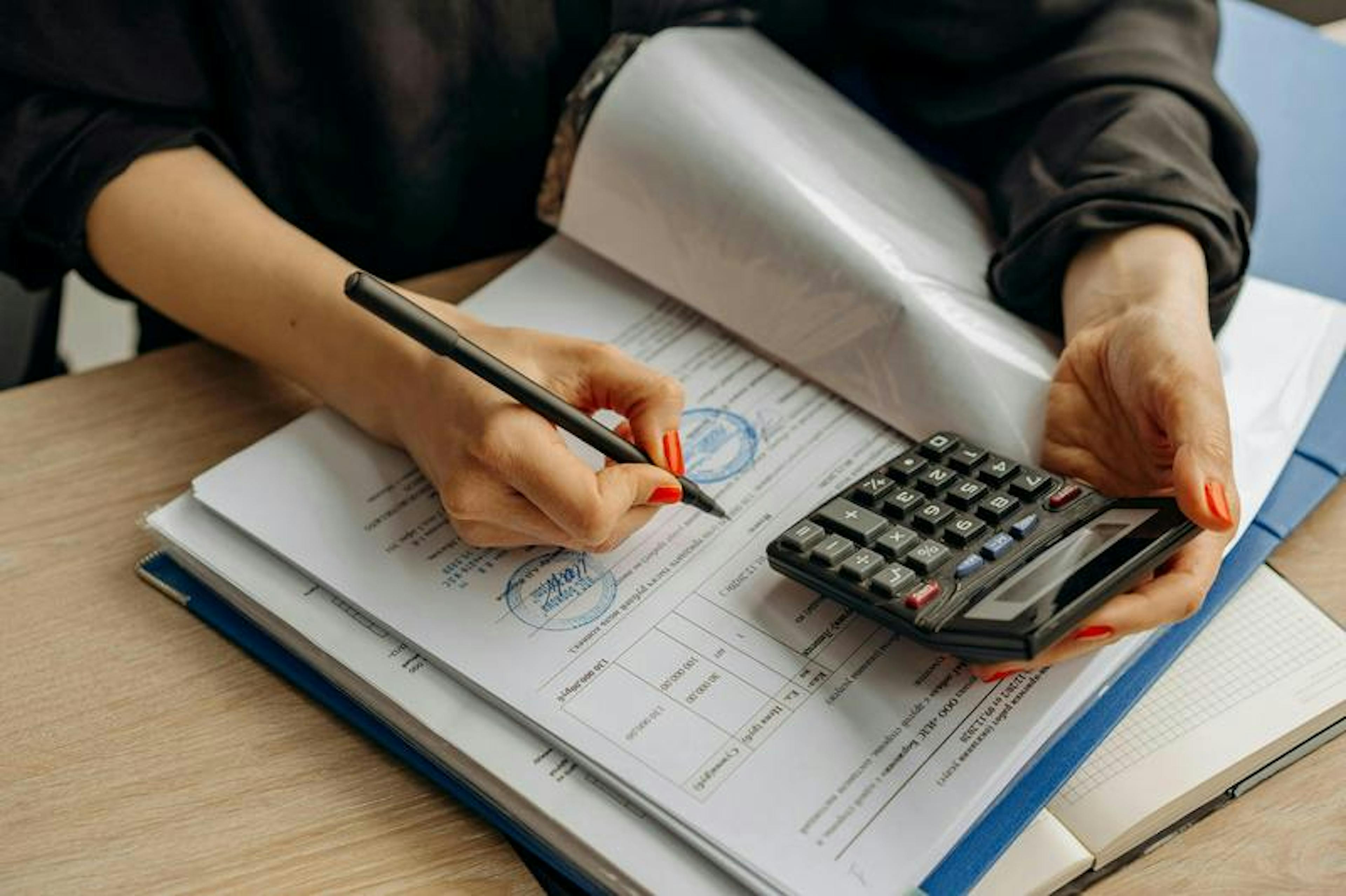 Person writes on documents with a pen while holding a calculator on a wooden desk in an office.
