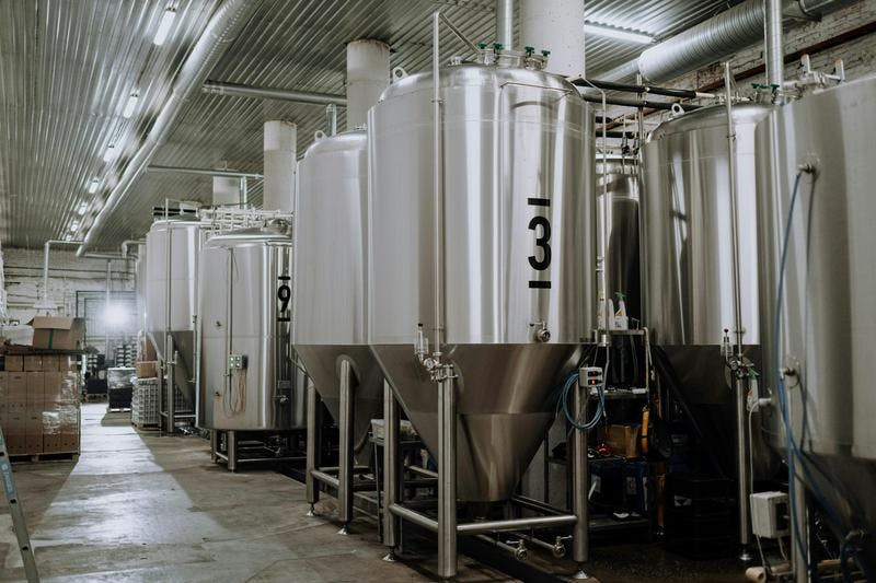 Large metal fermentation tanks are in an industrial brewery, with overhead lighting and pipes.