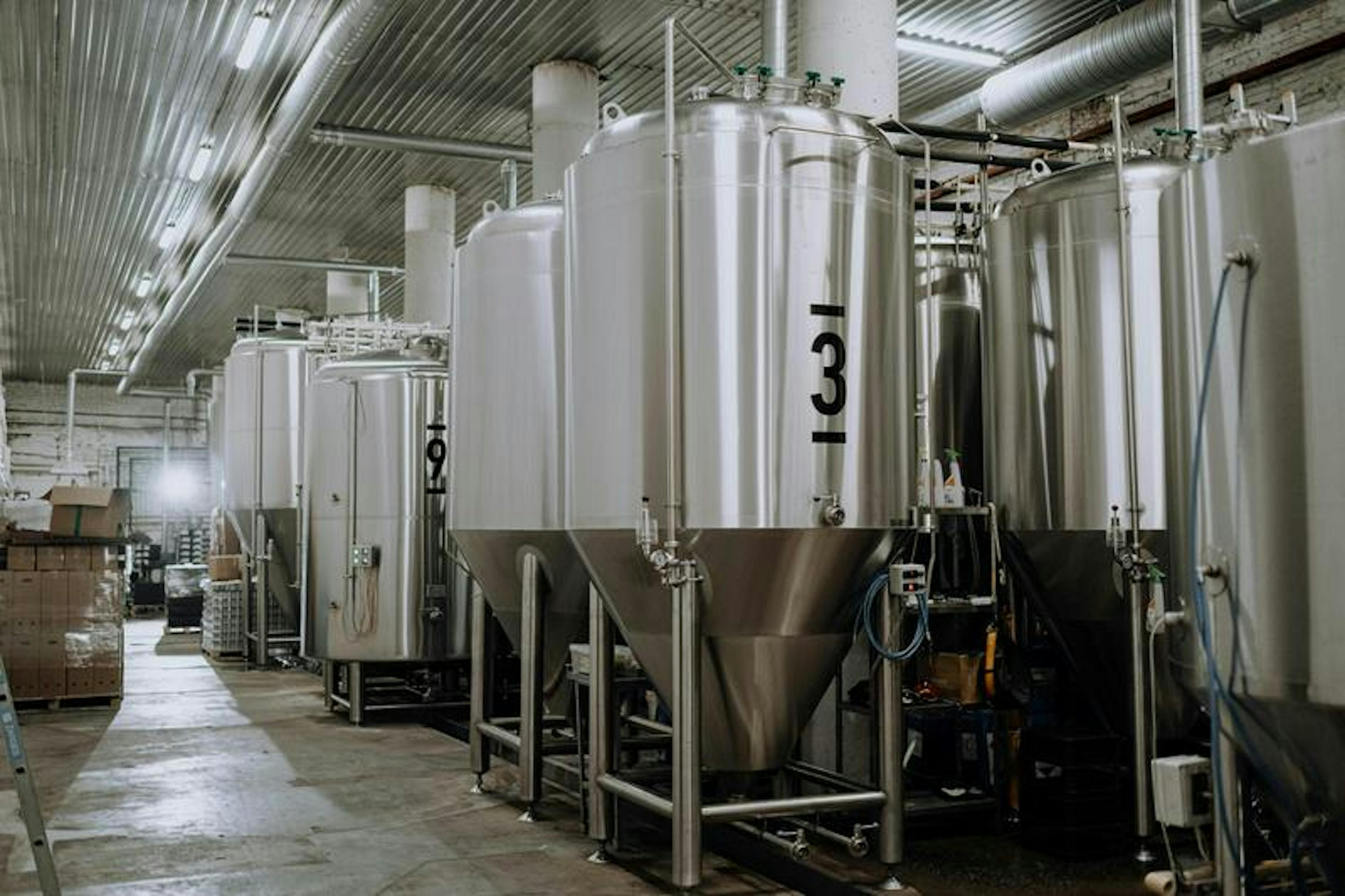 Large metal fermentation tanks are in an industrial brewery, with overhead lighting and pipes.