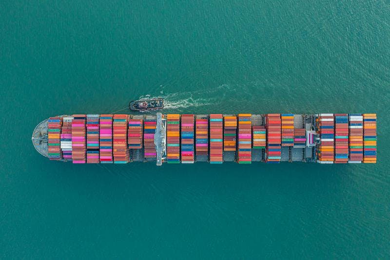 Large cargo ship with colorful containers towed by tugboat on calm ocean, viewed from above.