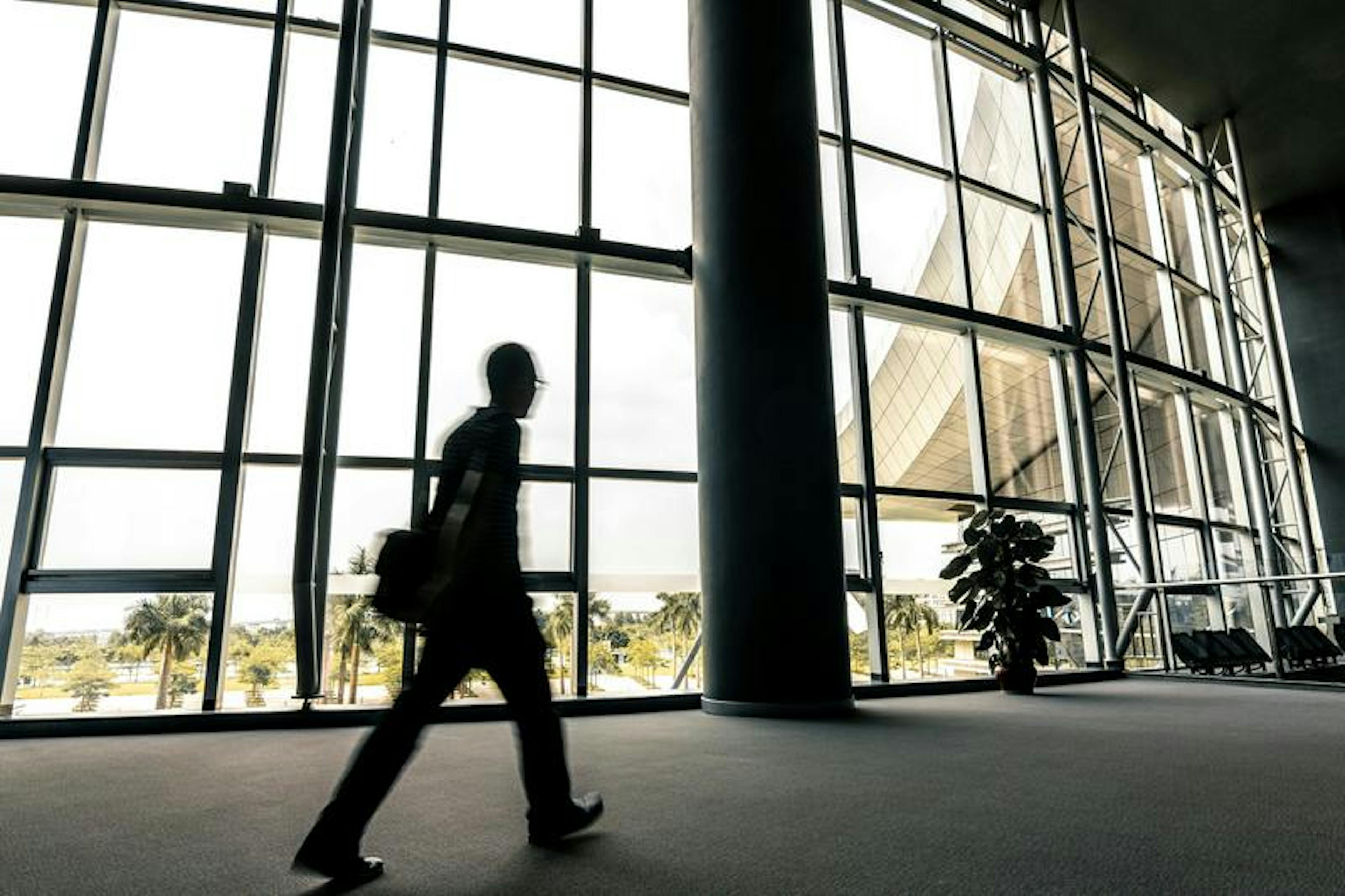 Silhouette of person walks past large glass windows; palm trees visible, enhancing spacious, sunny setting.