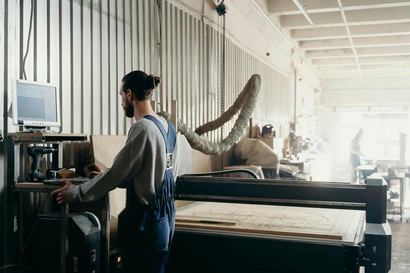 Person operates machinery in workshop, using computer to control CNC machine. Room with industrial equipment.