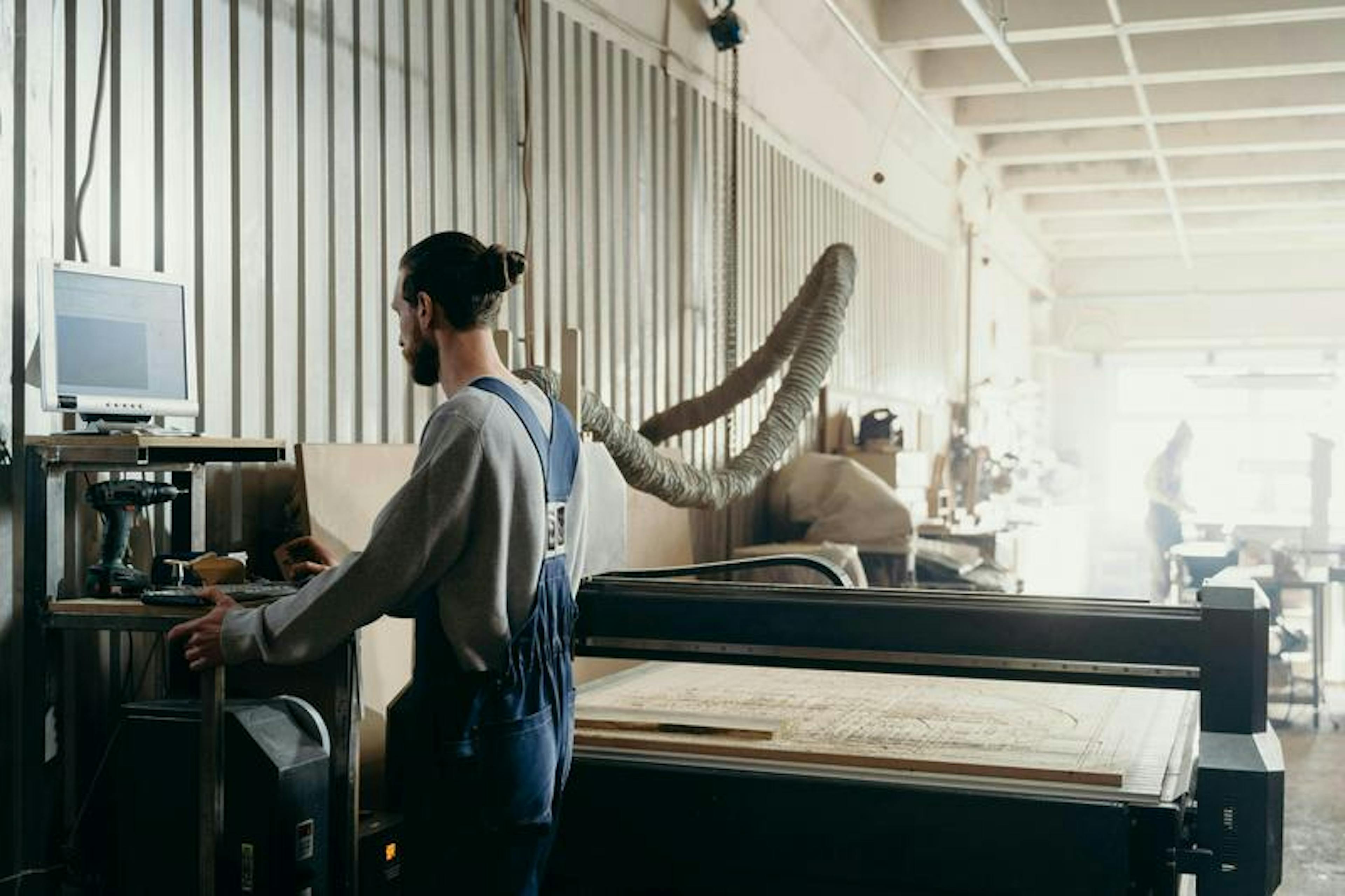 Person operates machinery in workshop, using computer to control CNC machine. Room with industrial equipment.