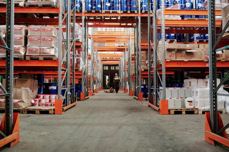 Worker pushes pallet jack with boxes down warehouse aisle, shelves filled with products, cans, cardboard packages.