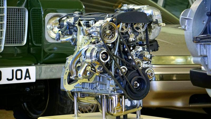 Intricate metallic car engine on stand in showroom; belts and pulleys visible, with two cars and "0 JOA" plate.