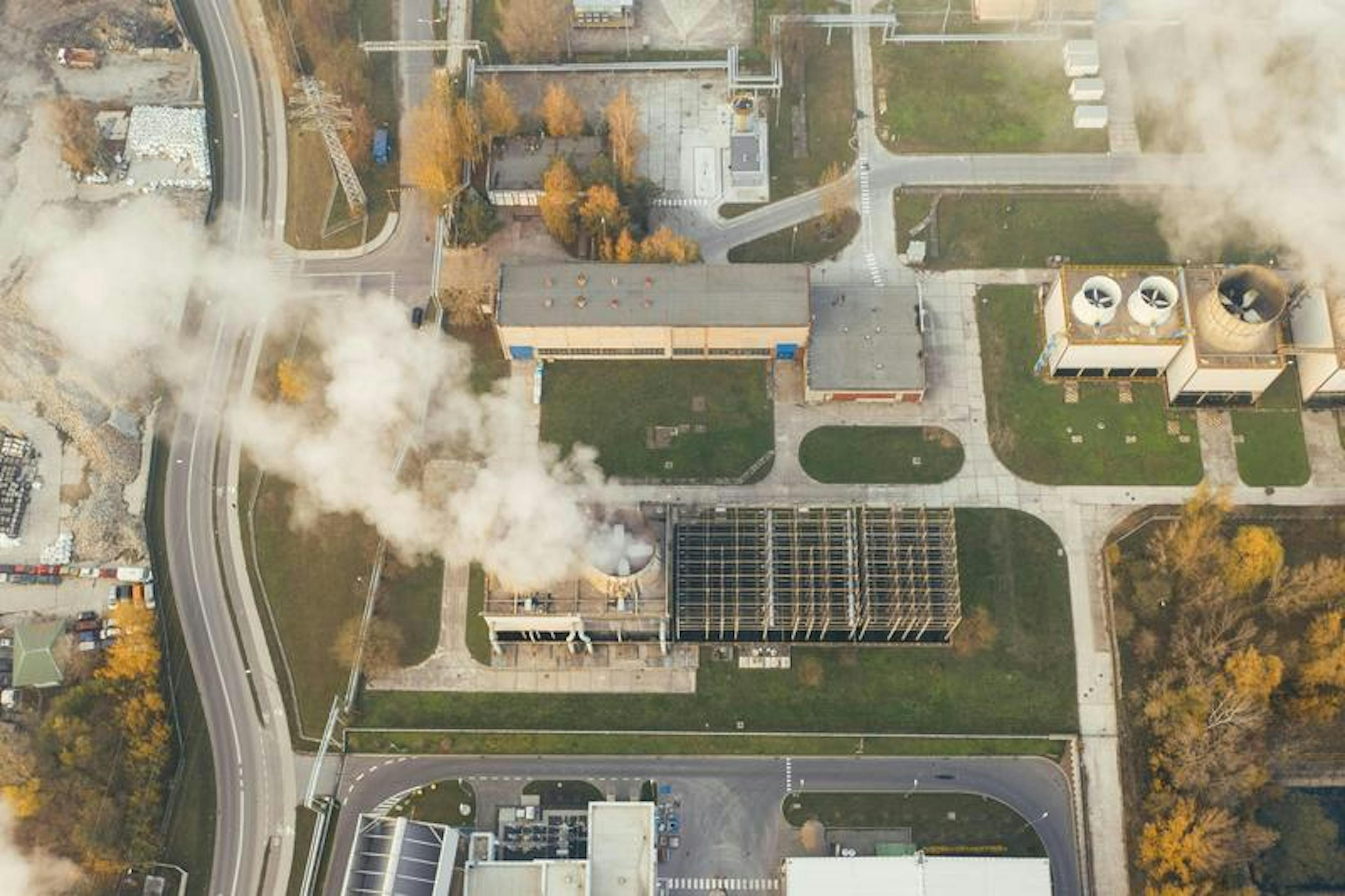 Smoke emerges from industrial chimneys, surrounded by buildings, roads, and autumn trees, in an aerial view.