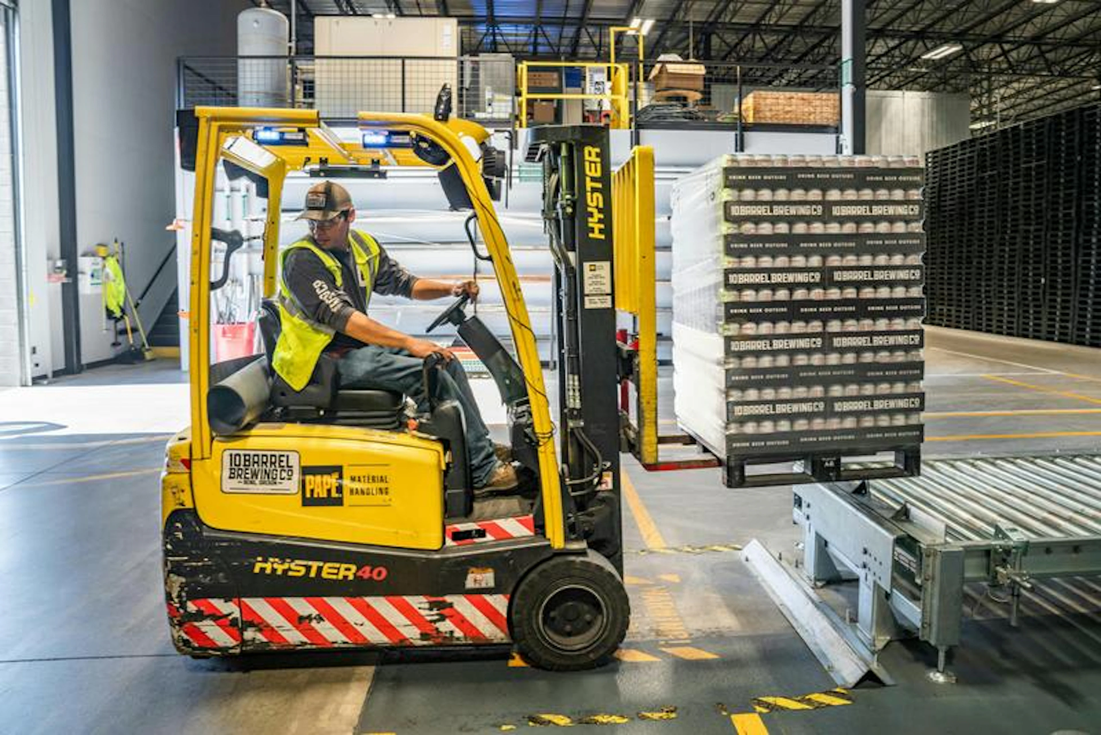 Forklift lifts 10 Barrel Brewing Co. boxes in warehouse. Signs: "PAPE MATERIAL HANDLING," "10 BARREL BREWING CO."